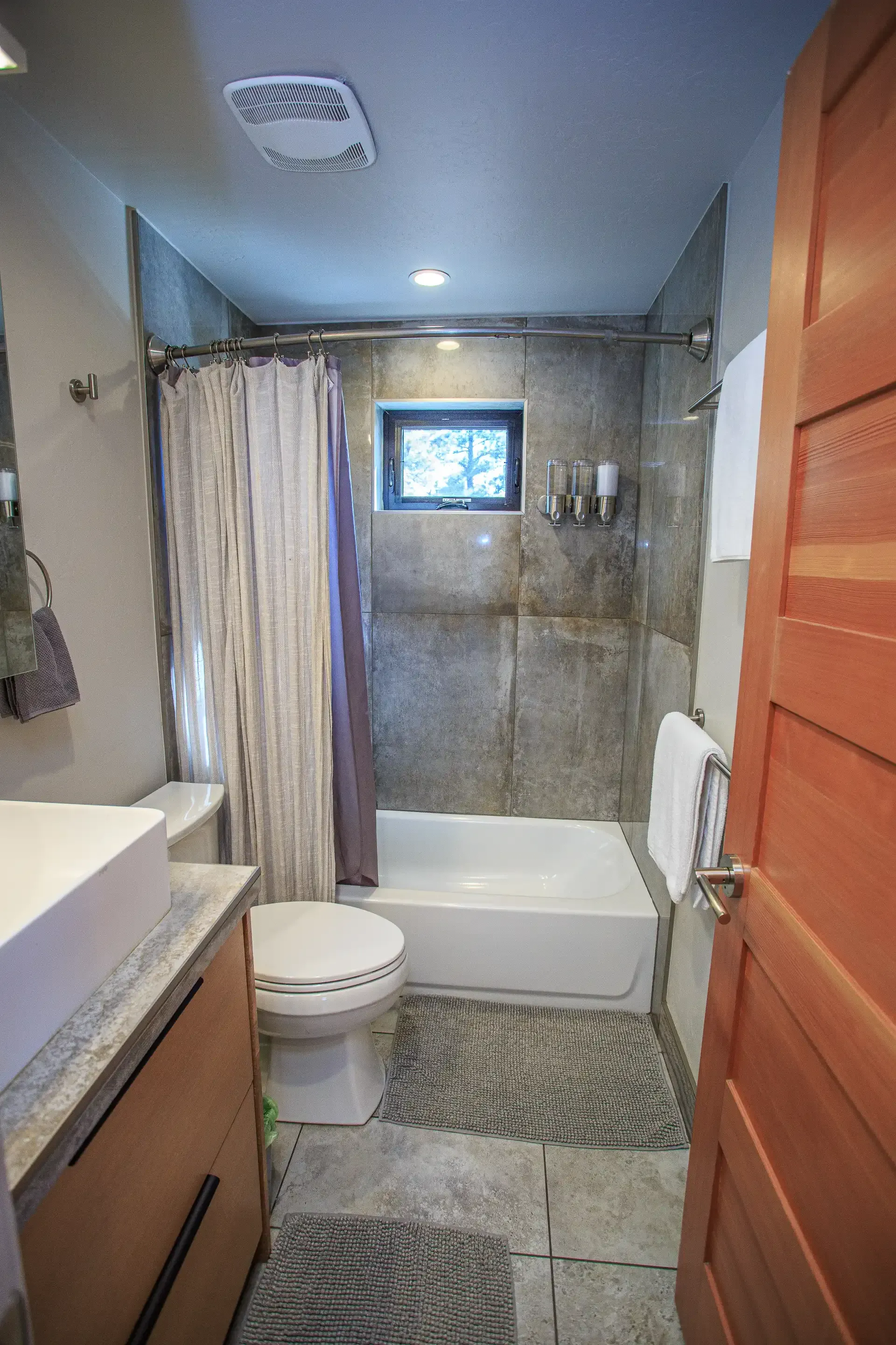 A bathroom featuring a light wood vanity, white toilet, and a bathtub with a shower curtain under a small window.