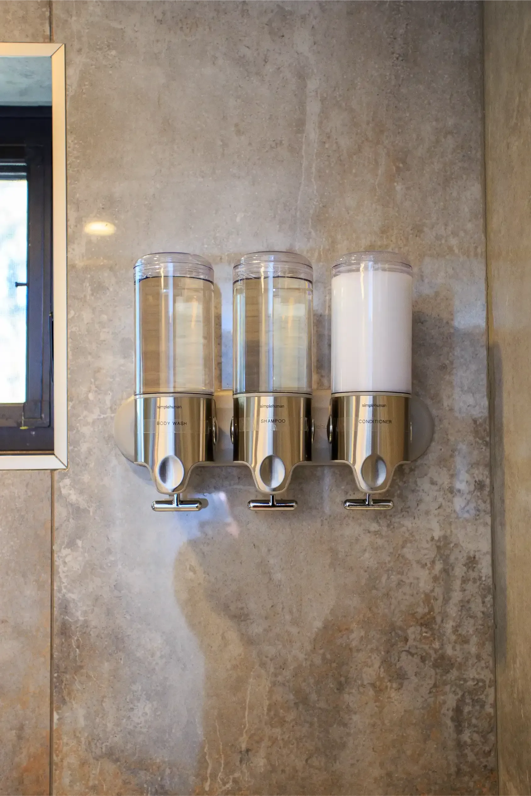 A three-chambered soap dispenser mounted on a textured, light brown stone wall.