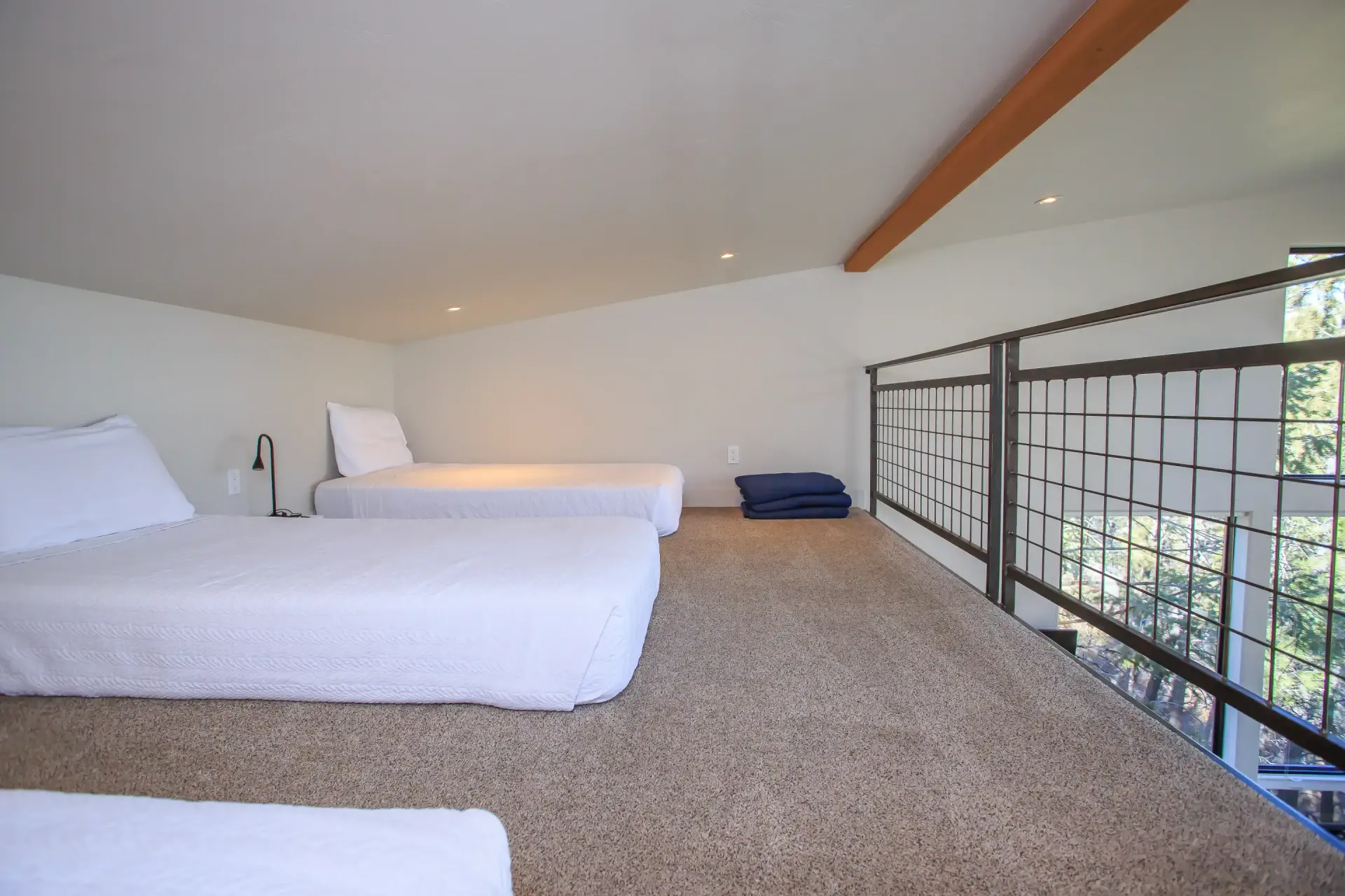 A carpeted bedroom loft with a sloping white ceiling, featuring three white beds and a metal safety railing.