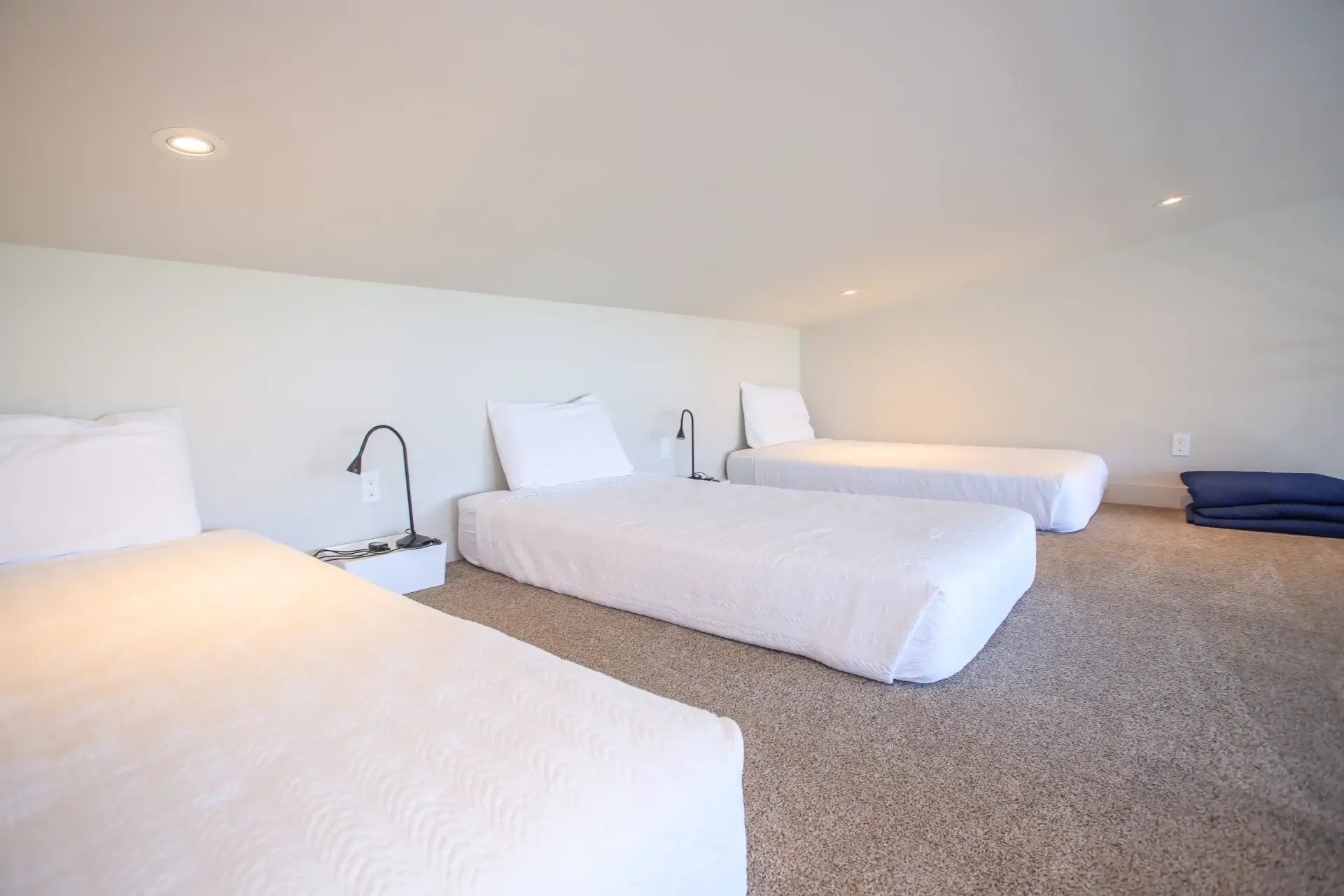 A bright, minimalist attic bedroom features three white single beds arranged on a carpeted floor under a sloped ceiling.