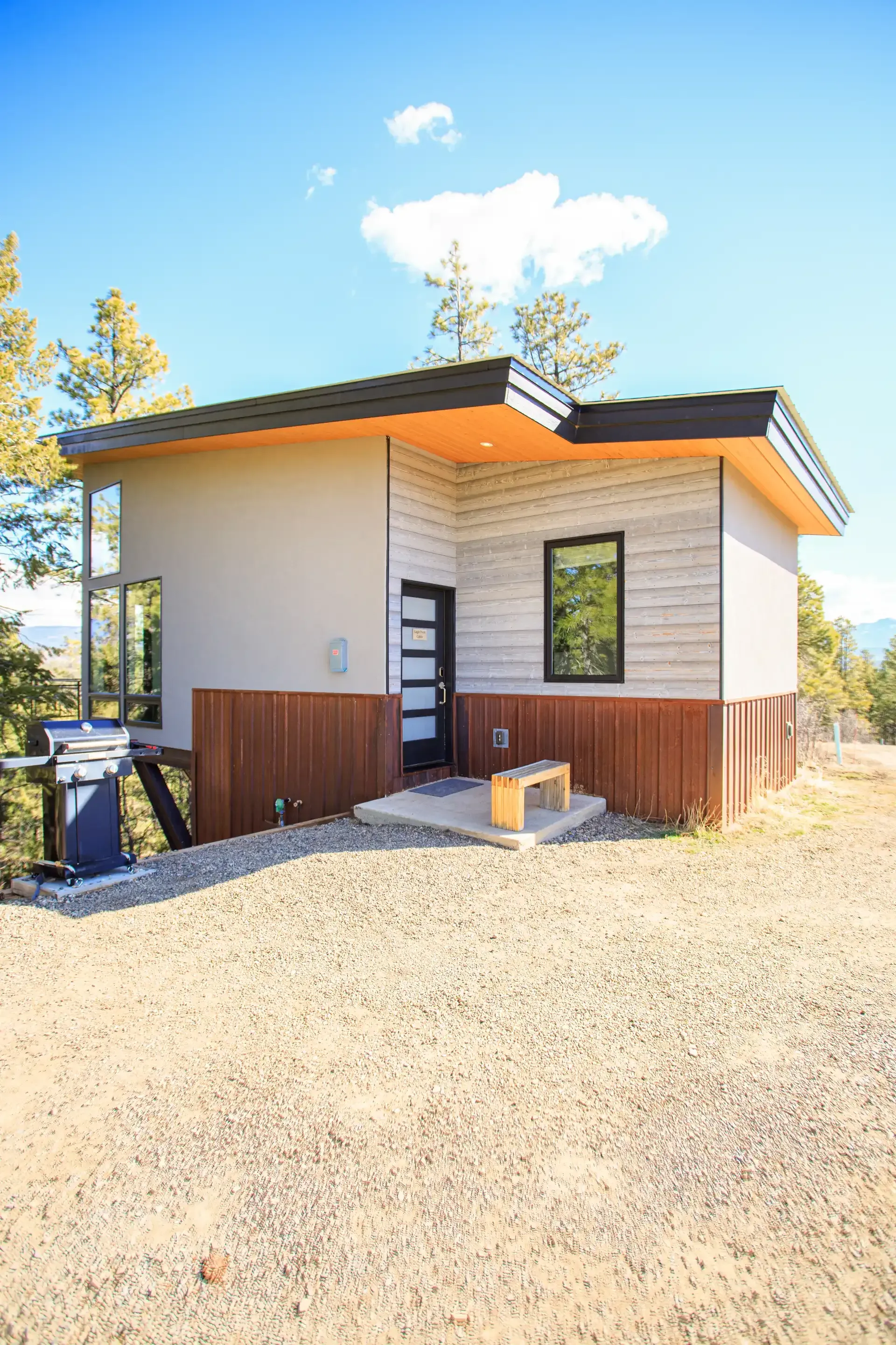 A modern cabin with tan and wood siding and a flat, overhanging roof stands on a gravel lot with trees under a blue sky.