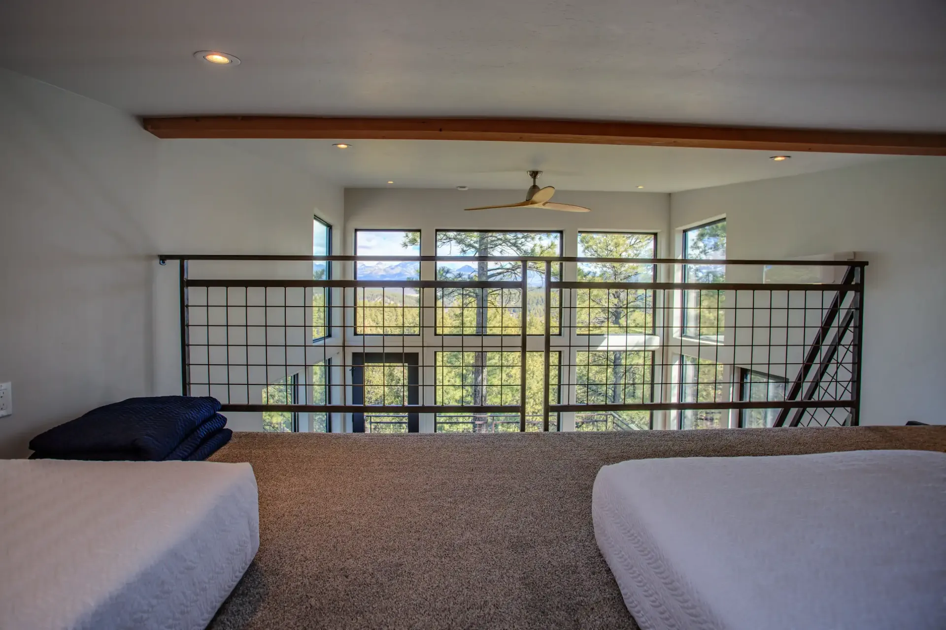 A carpeted loft space with two beds separated by a metal railing overlooking large windows with a forest view.