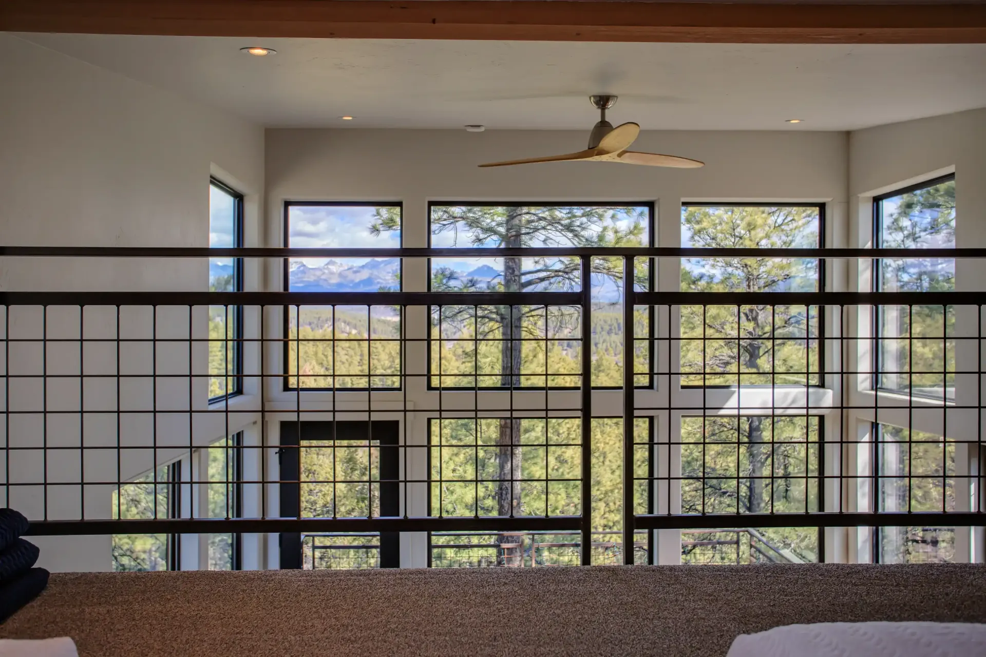A loft view overlooking a large living area with tall, gridded windows showing a forest and mountains in the distance.