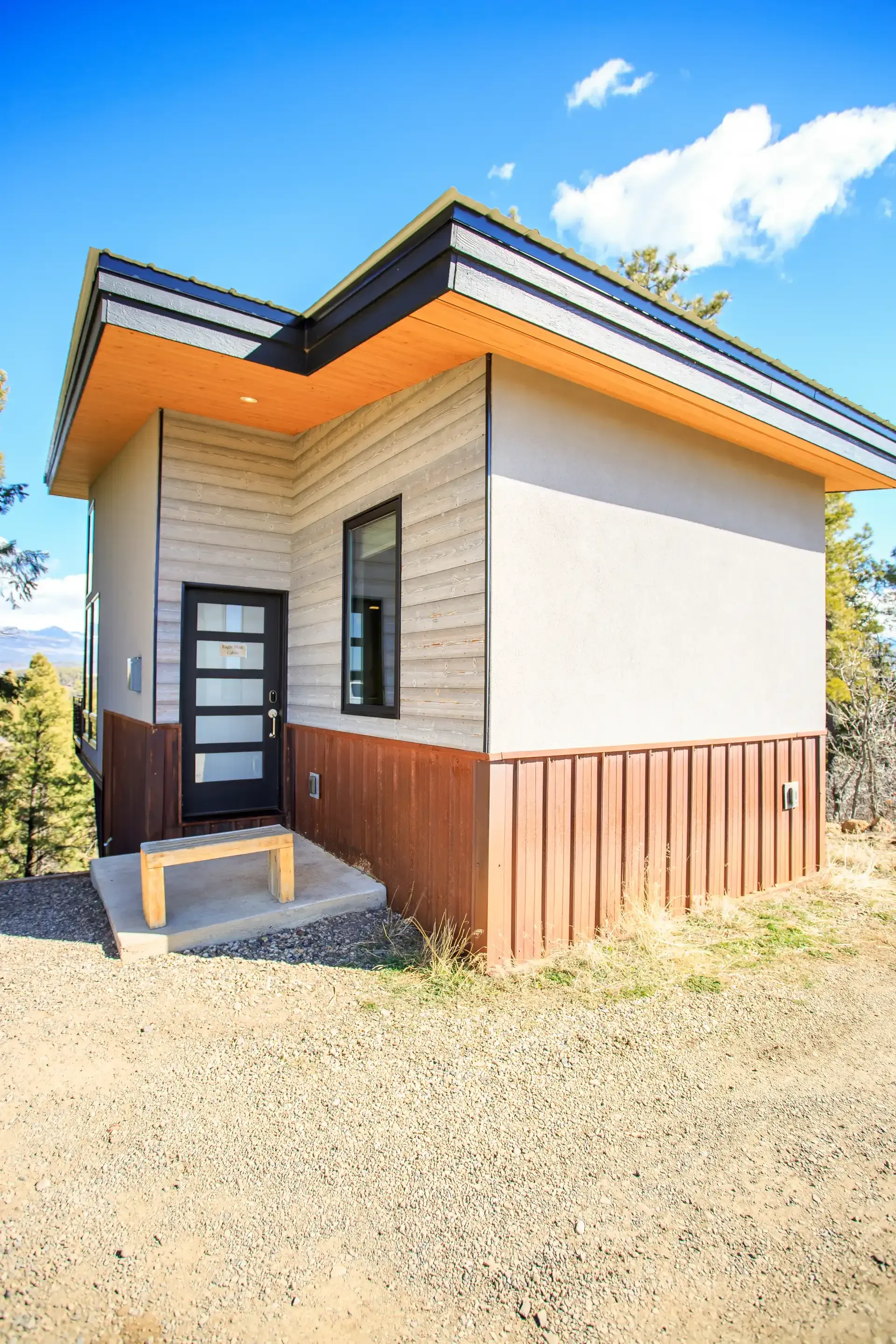 A modern, one-story cabin with horizontal siding, wood paneling, a flat roof, and a black front door on a gravel site.