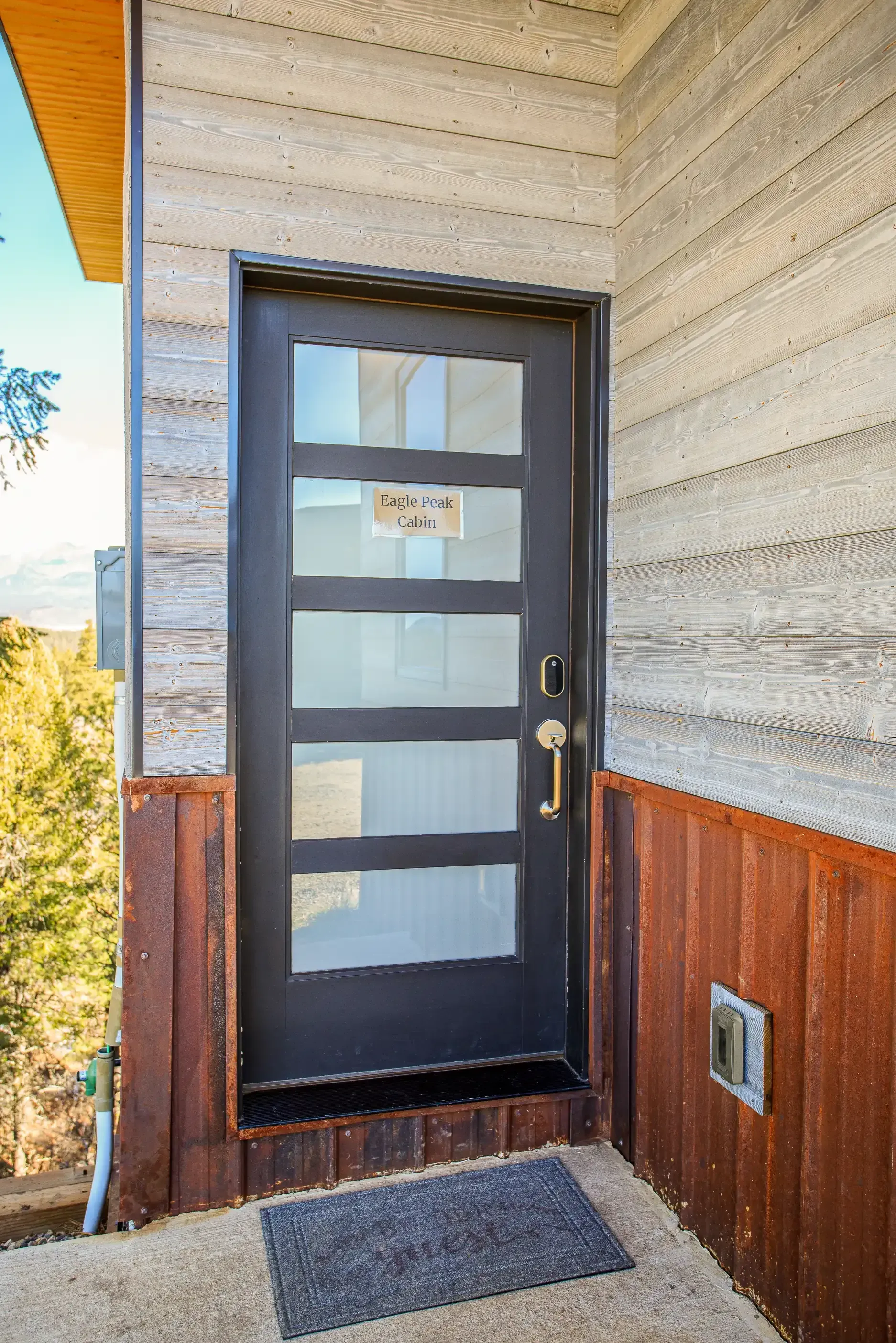 A dark door with five horizontal frosted glass panels set in a wall with light patterned siding and rusted metal trim.