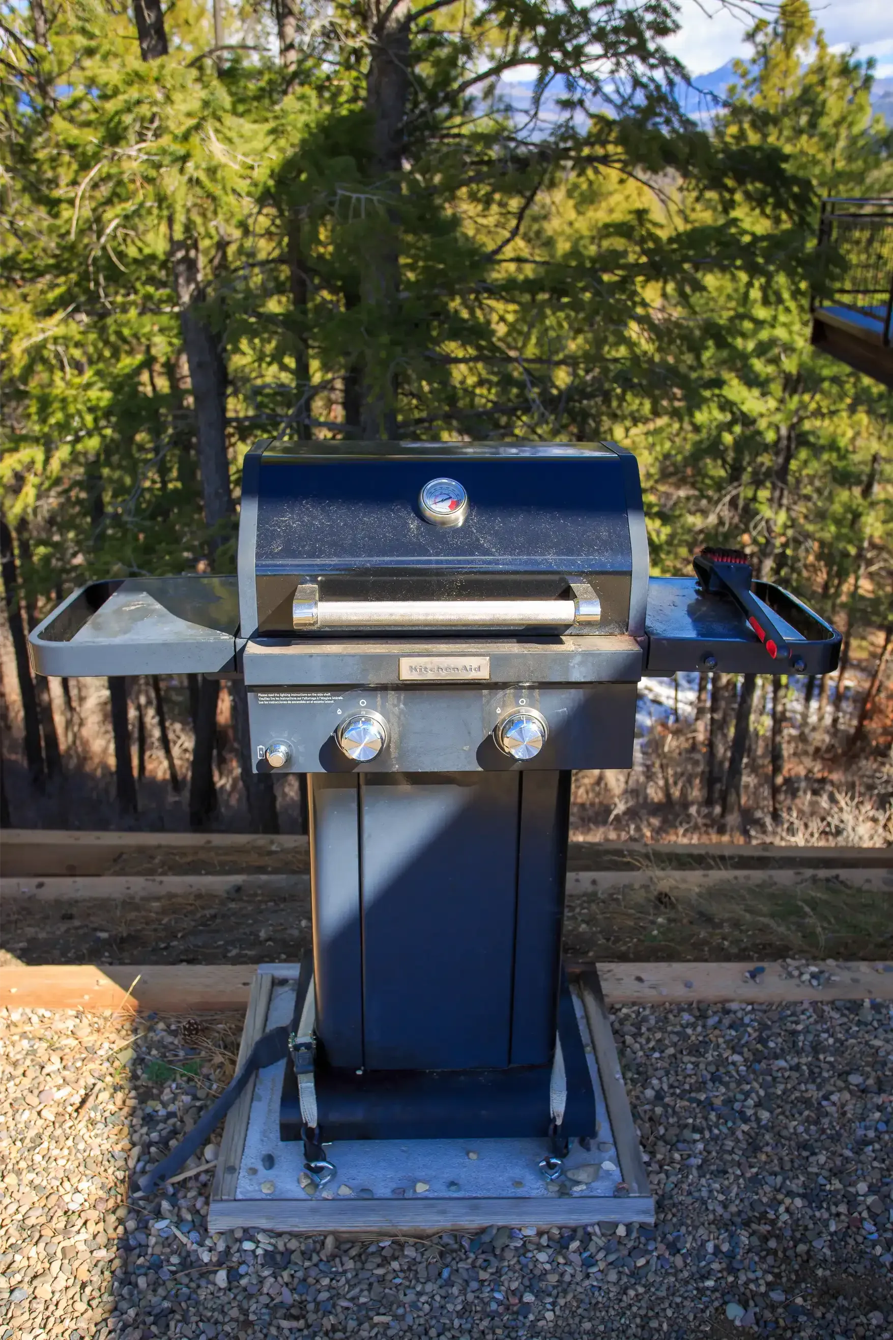 A dark gray two-burner gas grill with side shelves stands on a gravel patio outdoors against a background of trees.