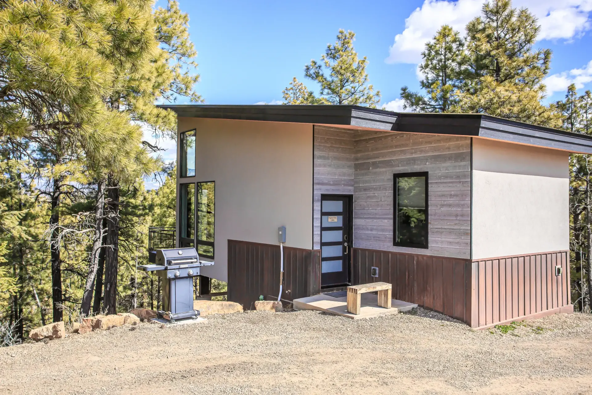 Modern cabin with wood and neutral siding, a front door, a grill, and a small bench set in a sunny, forested landscape.