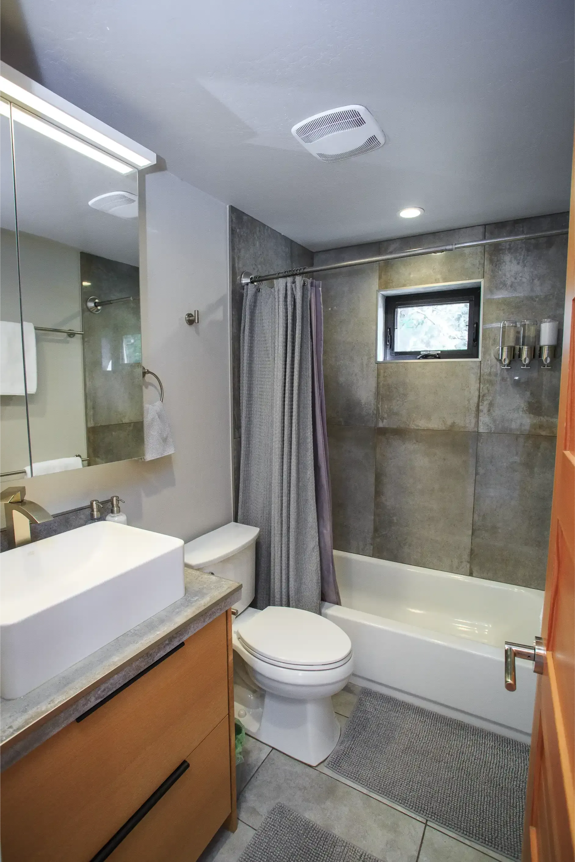 A modern bathroom with a wood vanity, white vessel sink, toilet, and a bathtub with a gray curtain and stone-look walls.