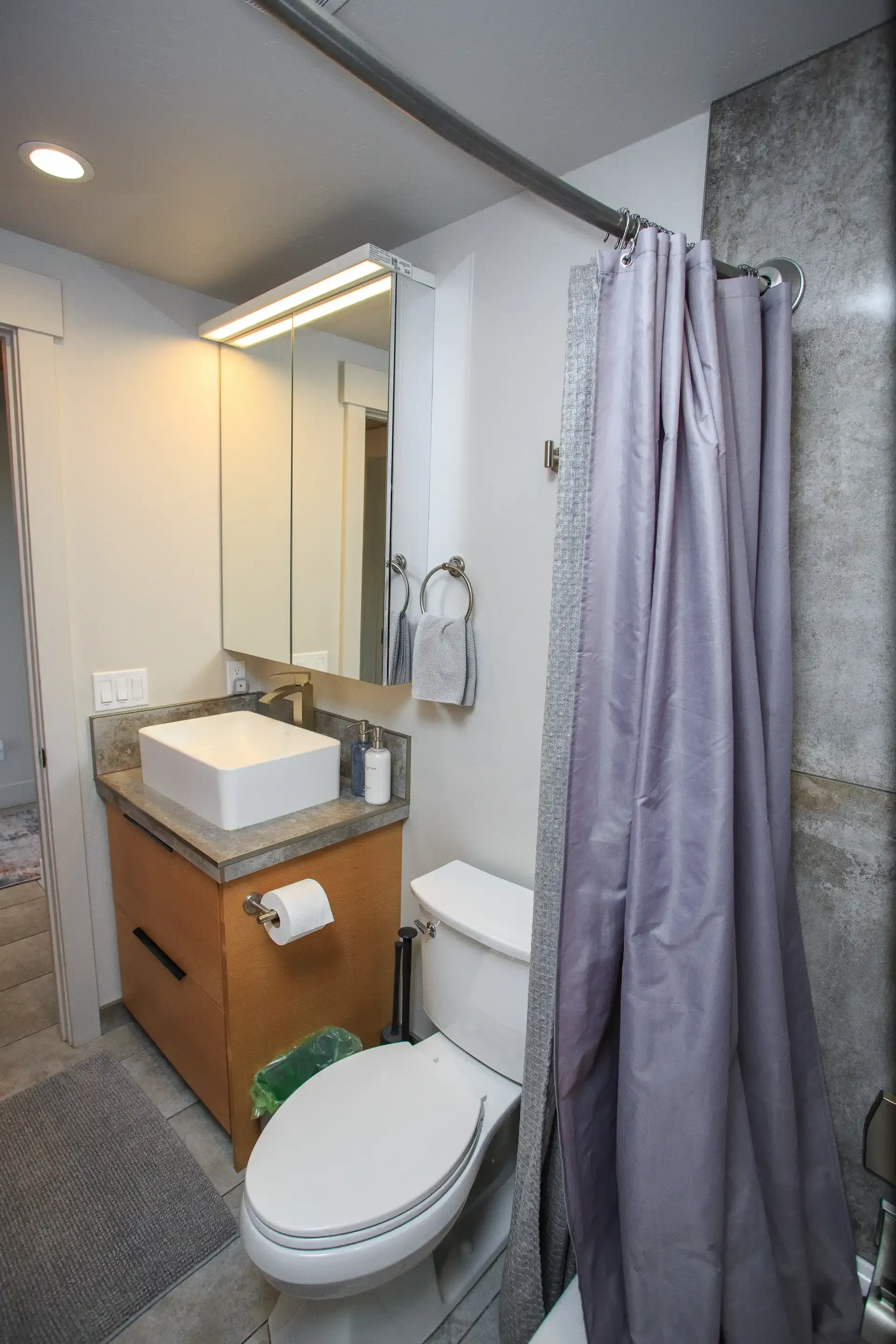 A small bathroom featuring a wooden vanity with a square sink, a toilet, and a shower with a lavender curtain.