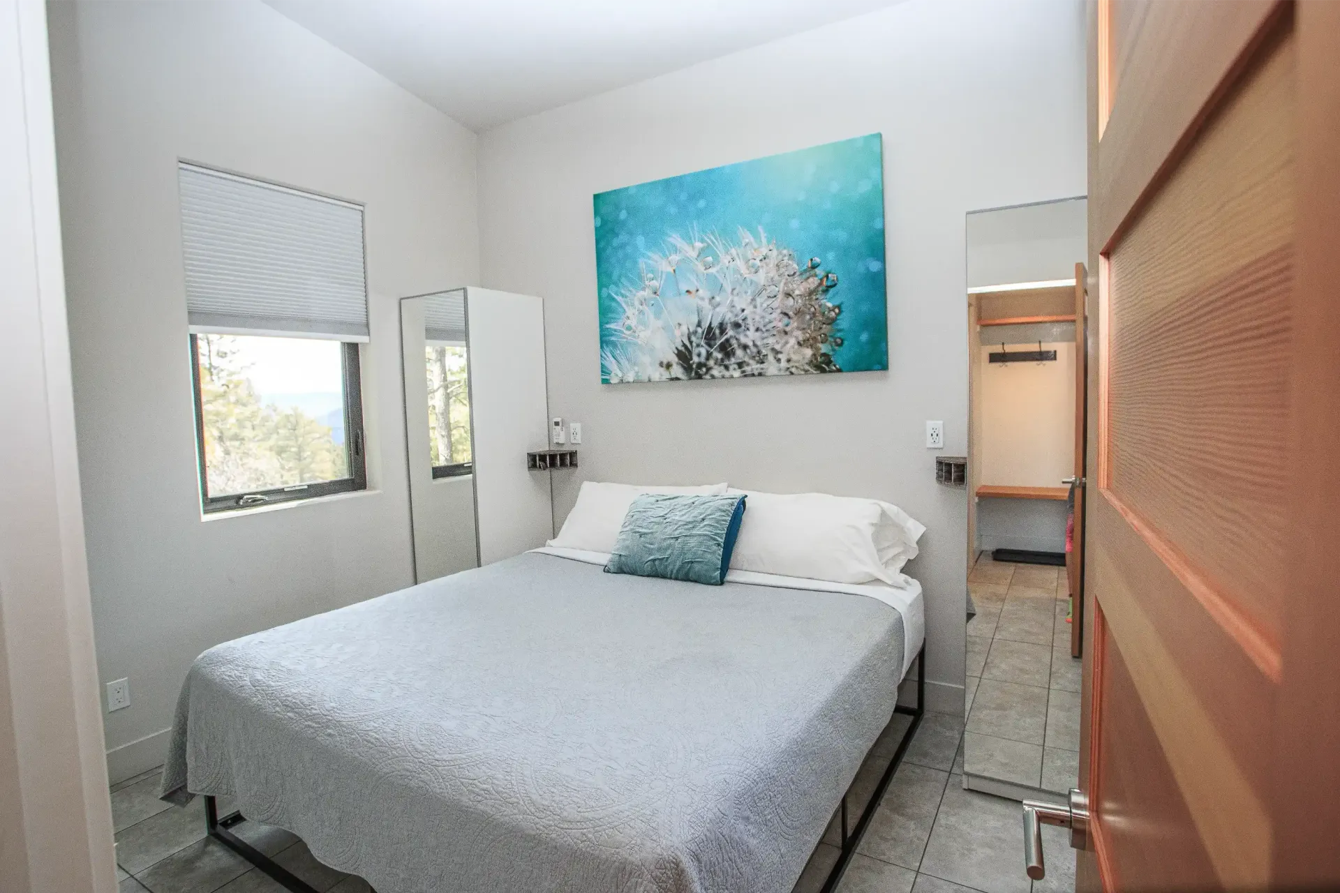 A bedroom with a queen bed, a gray quilt, a blue dandelion wall canvas, a window with blinds, and a mirrored closet.