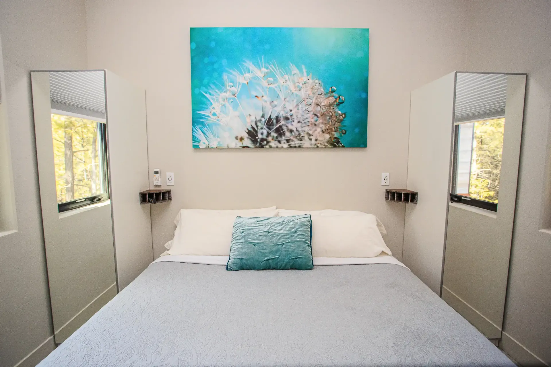 A bed with a blue textured pillow, white linens, and a large dandelion painting above it, flanked by two narrow mirrors.