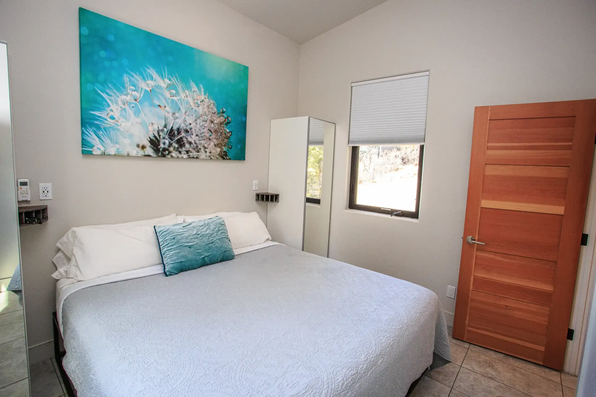 A bedroom with a bed featuring a gray quilt and blue pillow, a dandelion wall print, a window, and a wooden door.