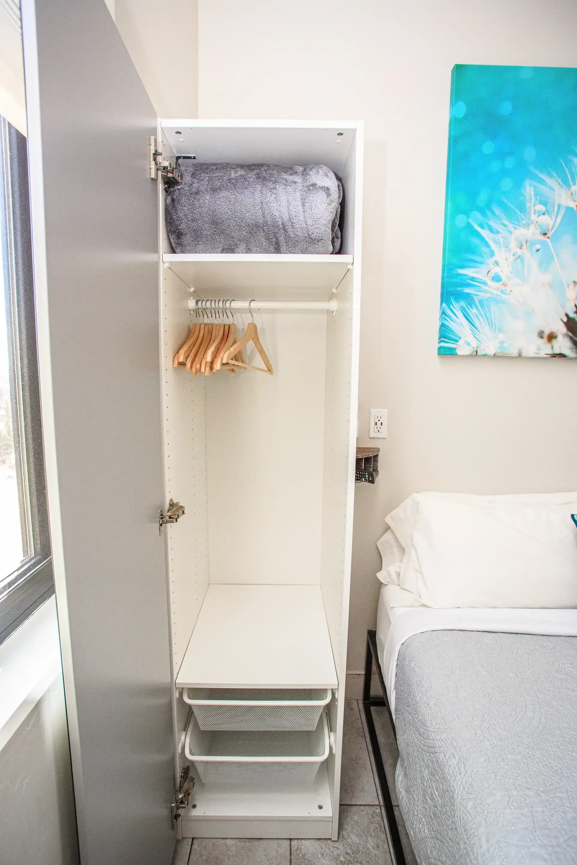 An open white wardrobe next to a bed, containing a grey blanket, wooden hangers, and two pull-out wire baskets.