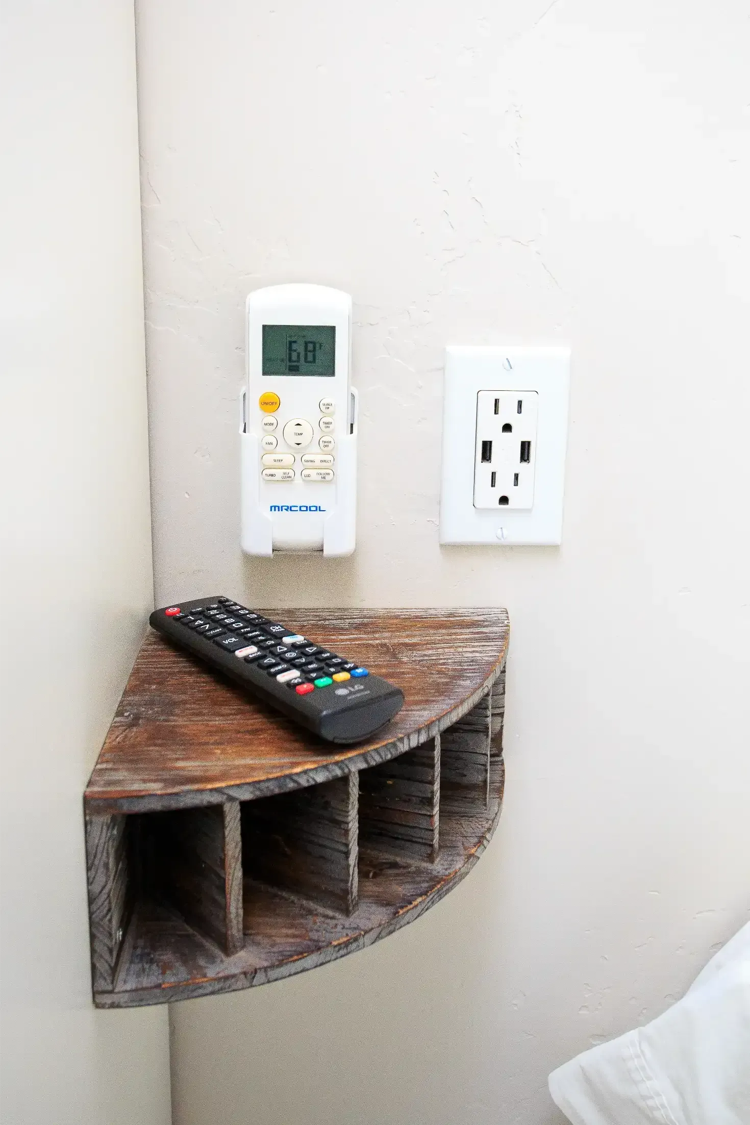 A dark wooden corner shelf holds a remote control, with an AC remote and a wall outlet mounted on the white wall above it.