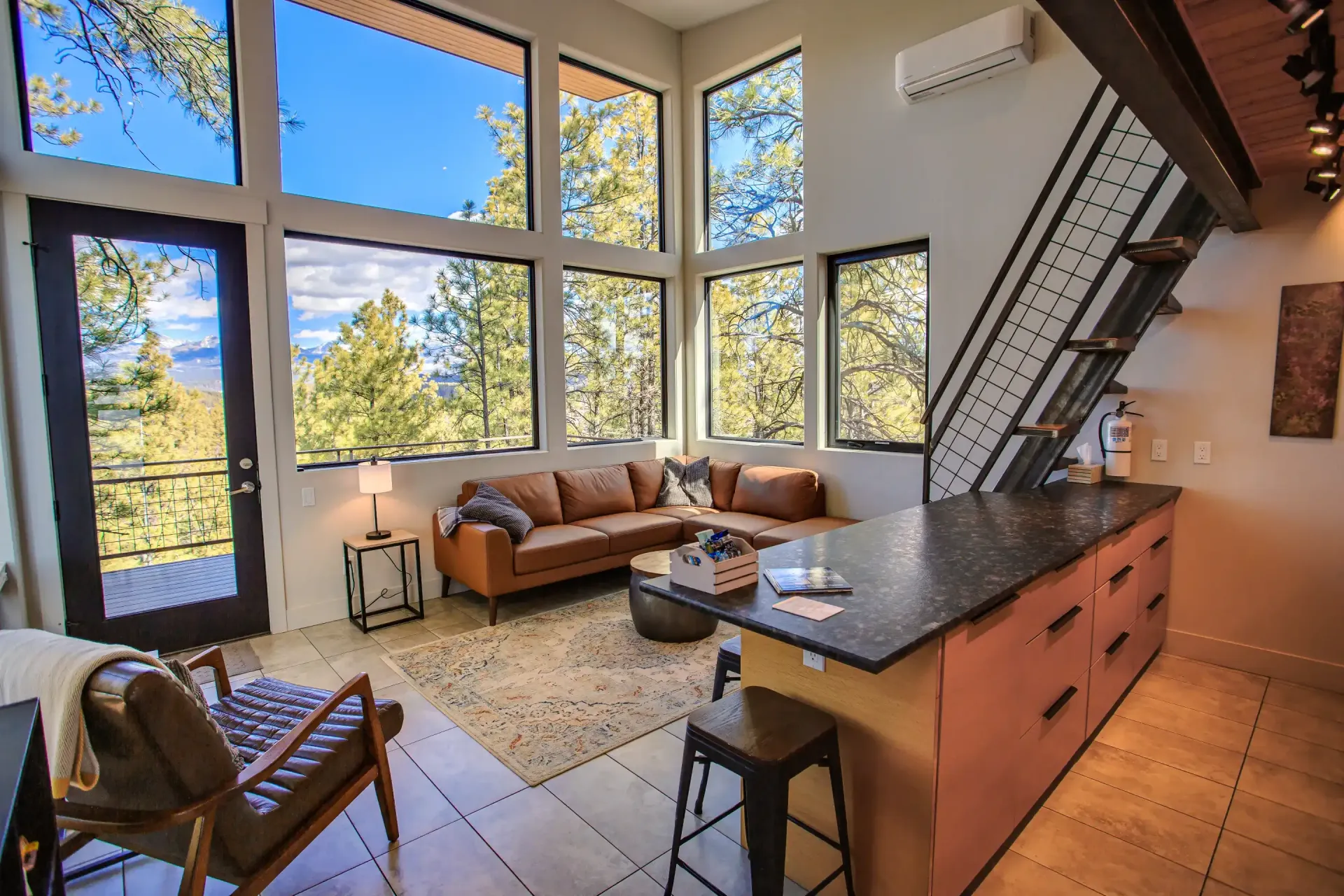 A cozy living room with large windows, a tan sectional sofa, leather chair, and a kitchen island with bar stools.
