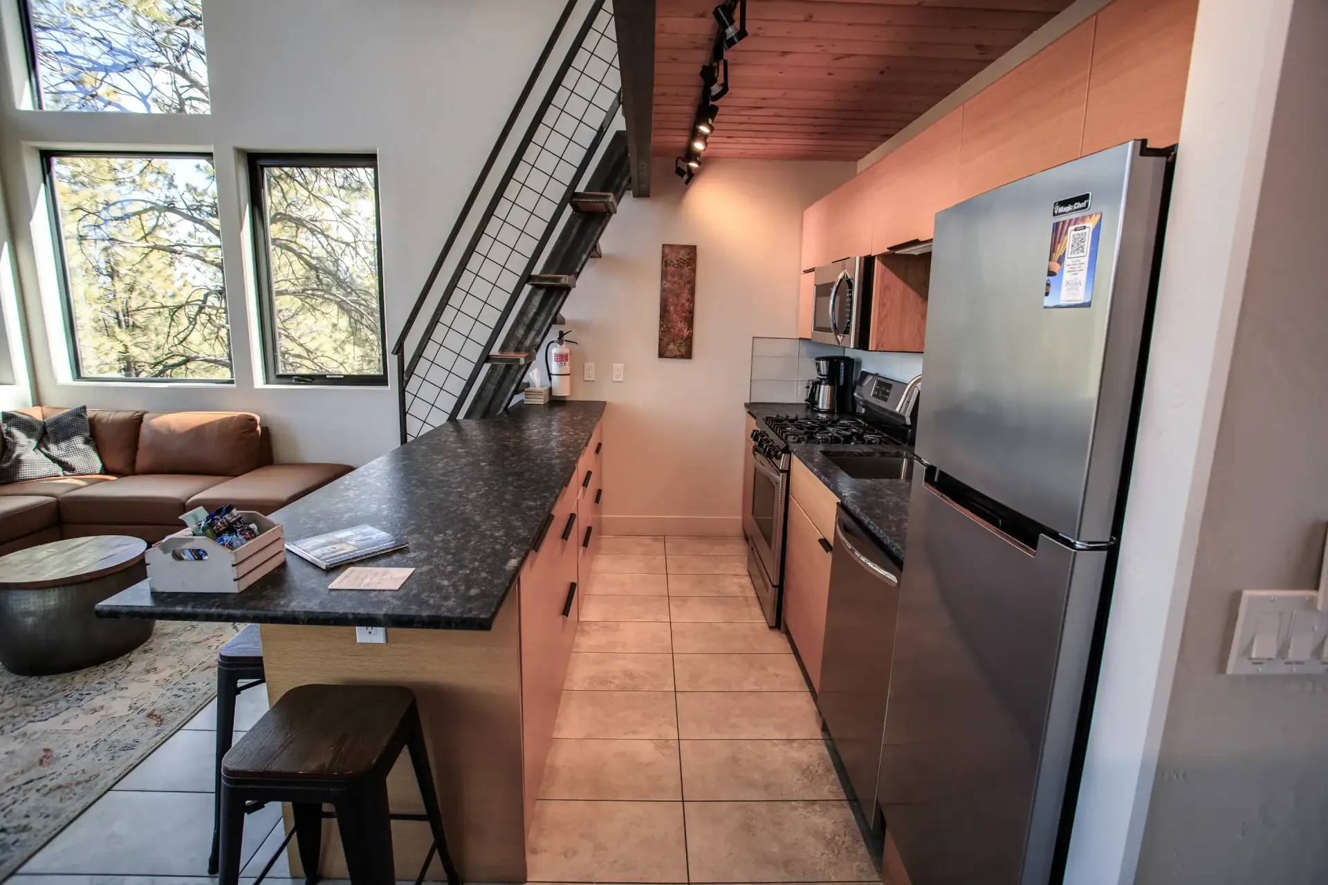 A modern kitchen with a black granite island, stainless steel appliances, and wood ceilings, next to a living area.