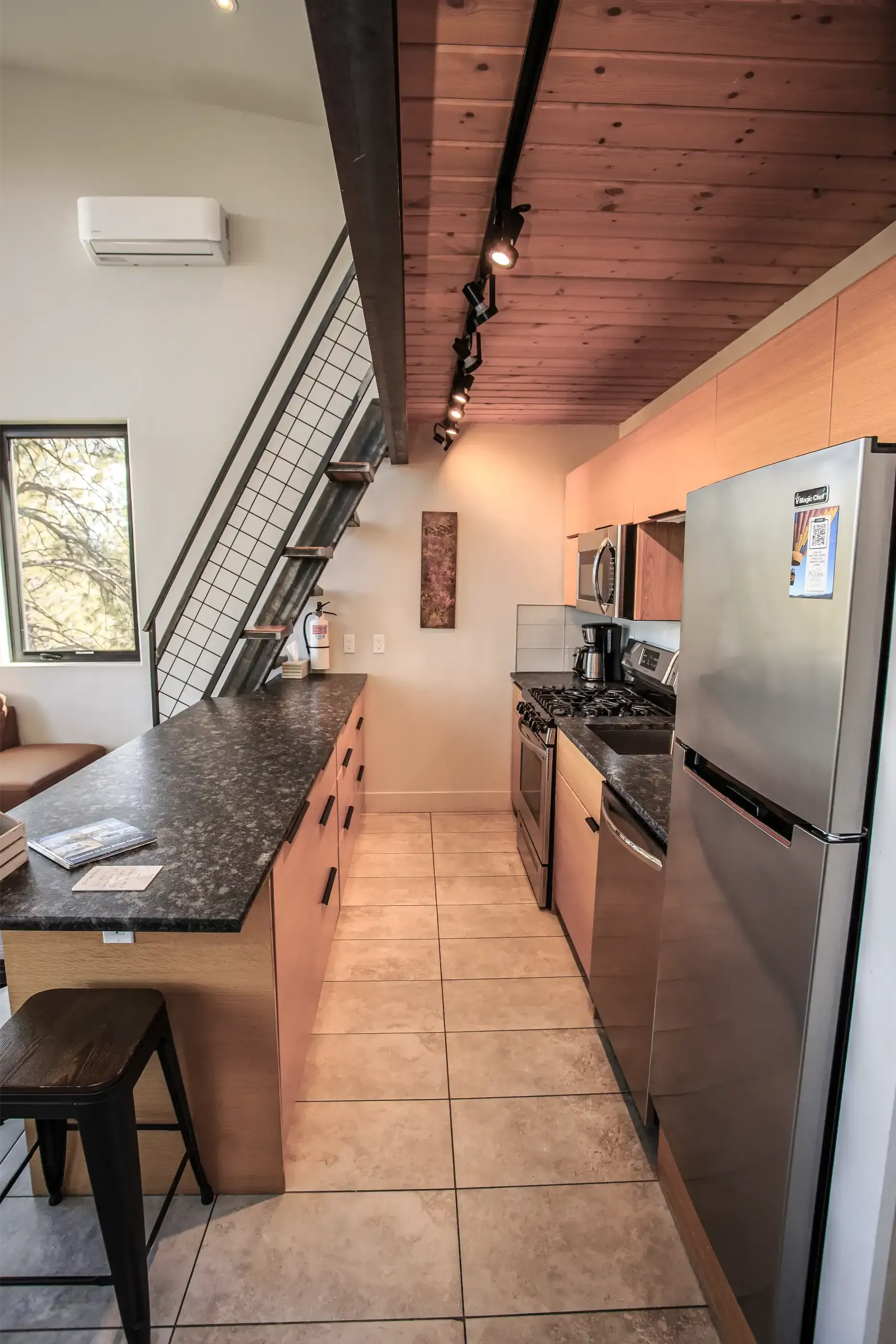 A kitchen with a long dark-speckled island, stainless steel appliances, light wood cabinets, and a wood-paneled ceiling.