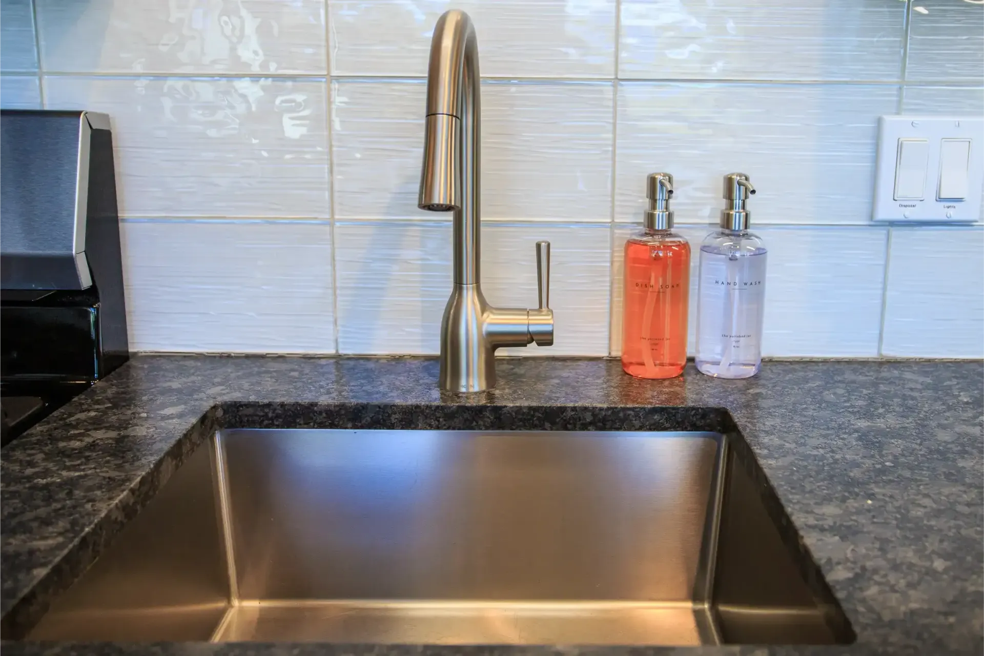 A kitchen sink with a stainless steel faucet and two soap dispensers sitting on a dark granite countertop.