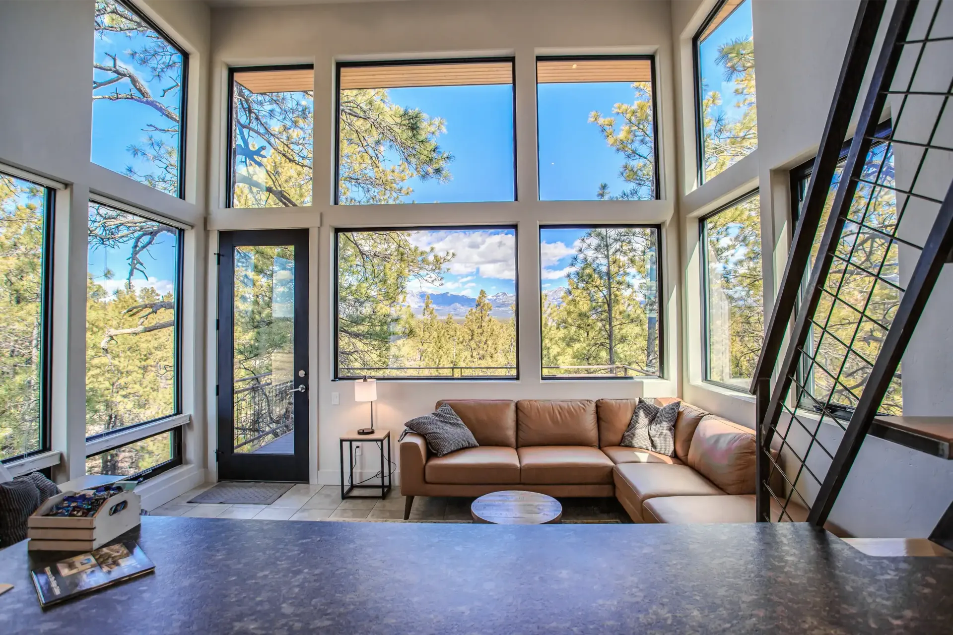 Living room with a tan leather sectional sofa, floor-to-ceiling windows looking out on trees, and a dark metal staircase.
