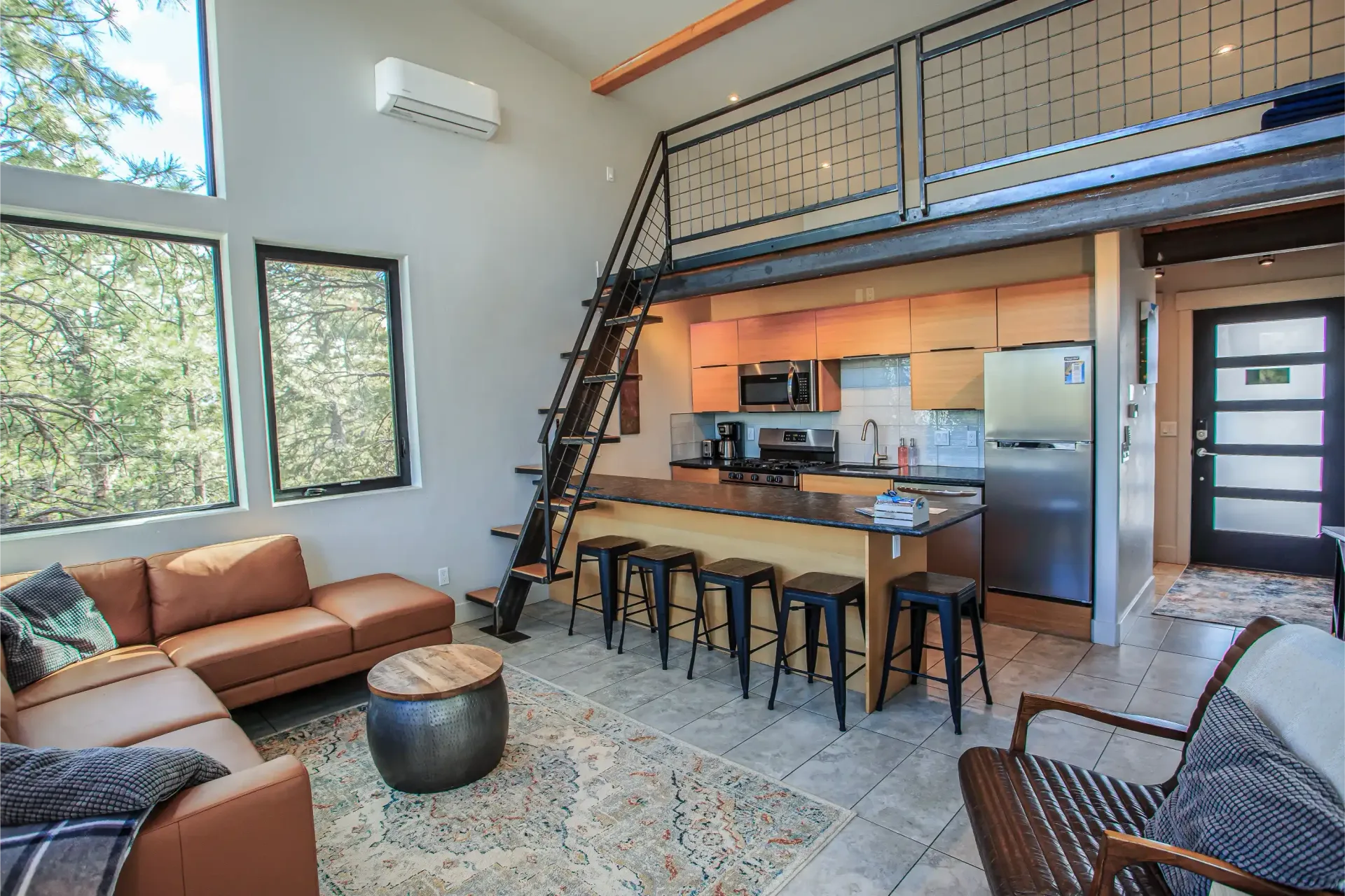 A modern open-plan living area with a tan leather sofa, kitchen island with bar stools, loft space, and large windows.