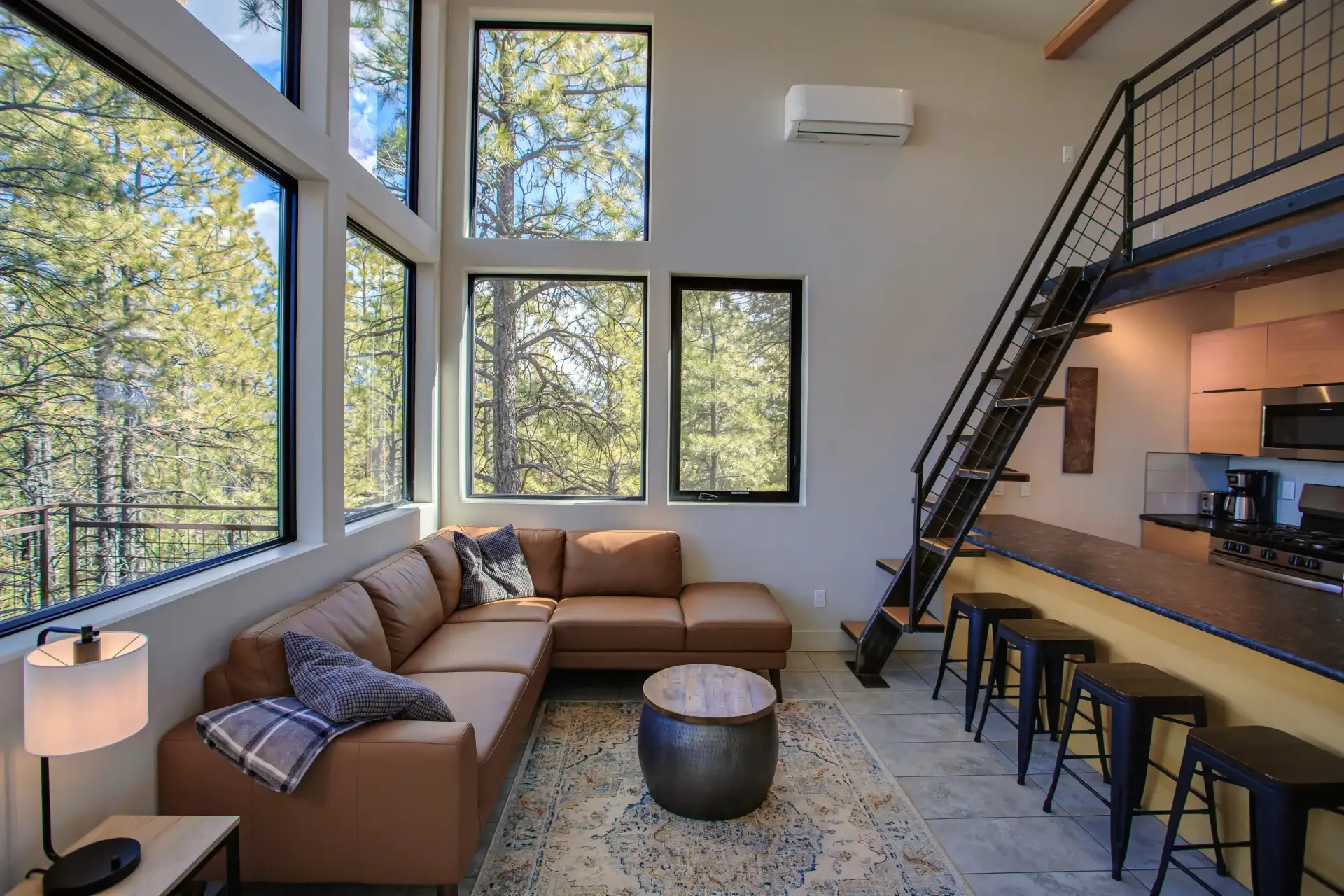 Modern living room with a brown sectional, floor-to-ceiling windows, wooden stairs, and a kitchen bar with stools.