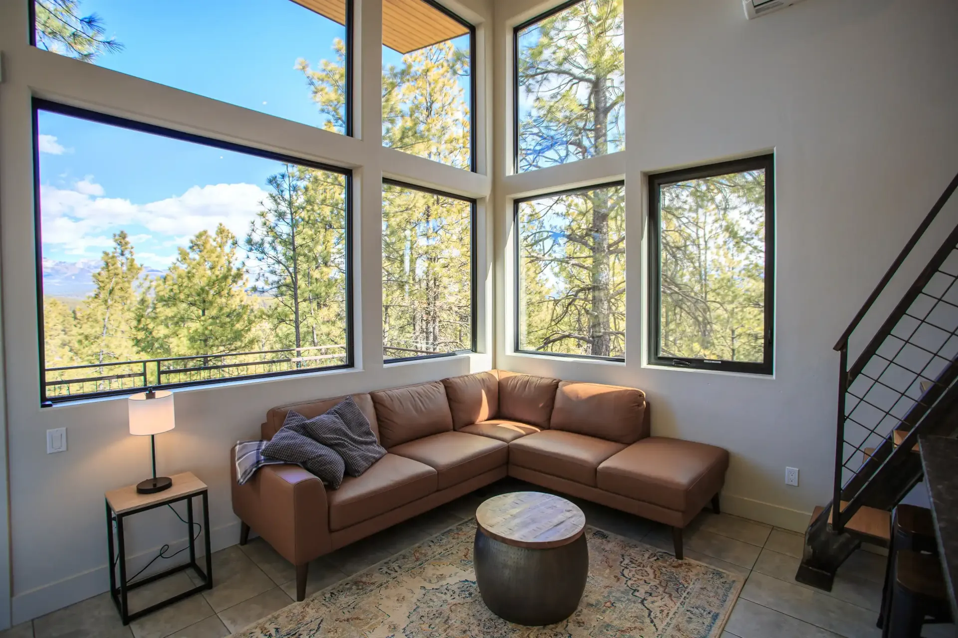 A modern living room with a brown sectional sofa and circular ottoman under large windows overlooking a forest.