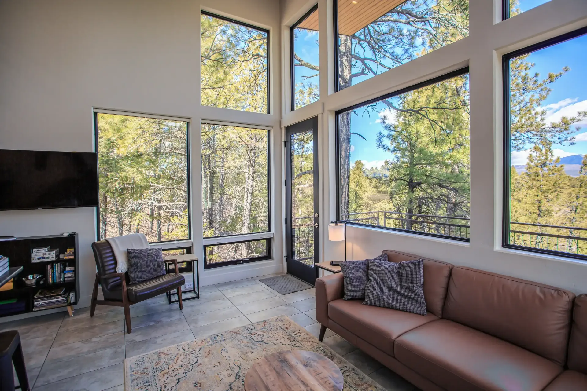 Modern living room with floor-to-ceiling windows, leather sofa, armchair, and TV, overlooking a pine forest.