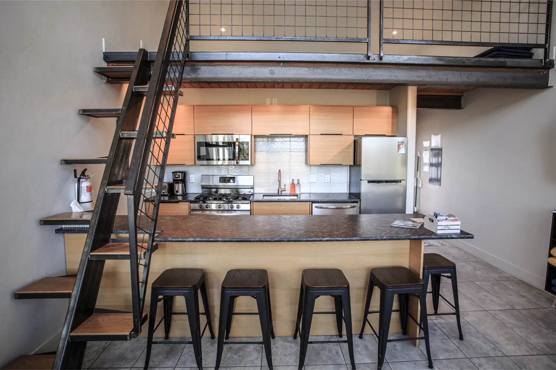 A kitchen with light wood cabinets, stainless appliances, a granite bar, four dark stools, and a metal ladder to a loft.
