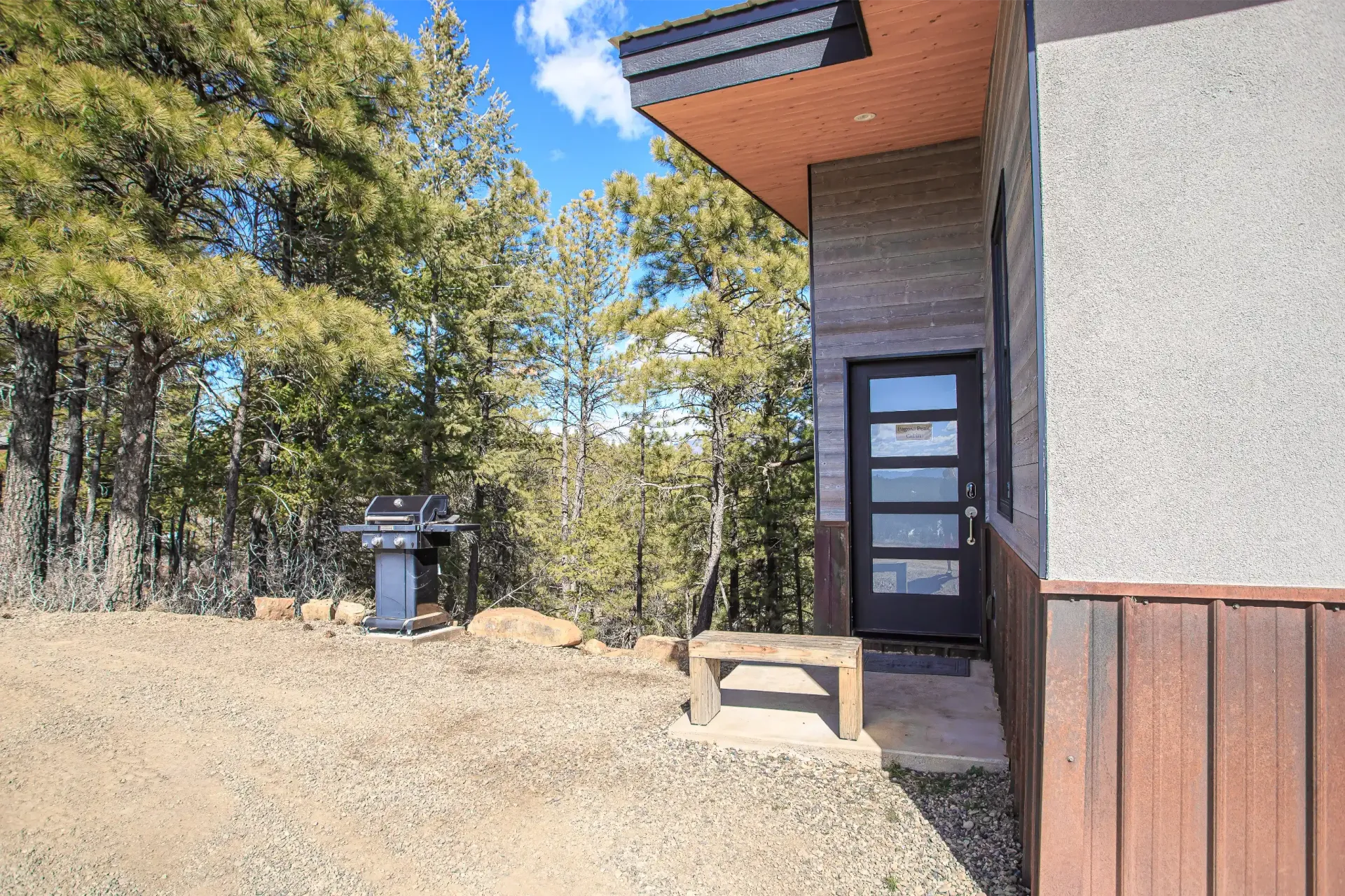 A brown modern house exterior with a glass door, a small stone bench, and a charcoal grill on a dirt lot near pine trees.