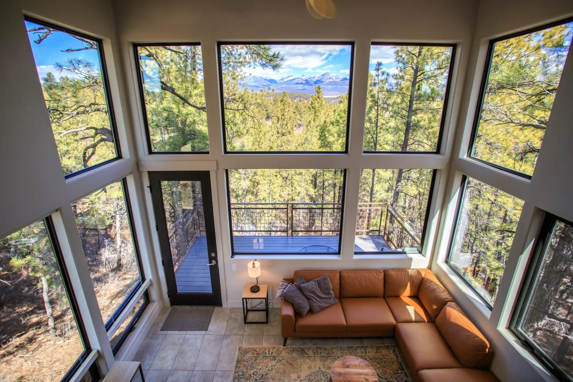 Modern living room with a large leather sectional, wood floors, and floor-to-ceiling windows overlooking a forest.