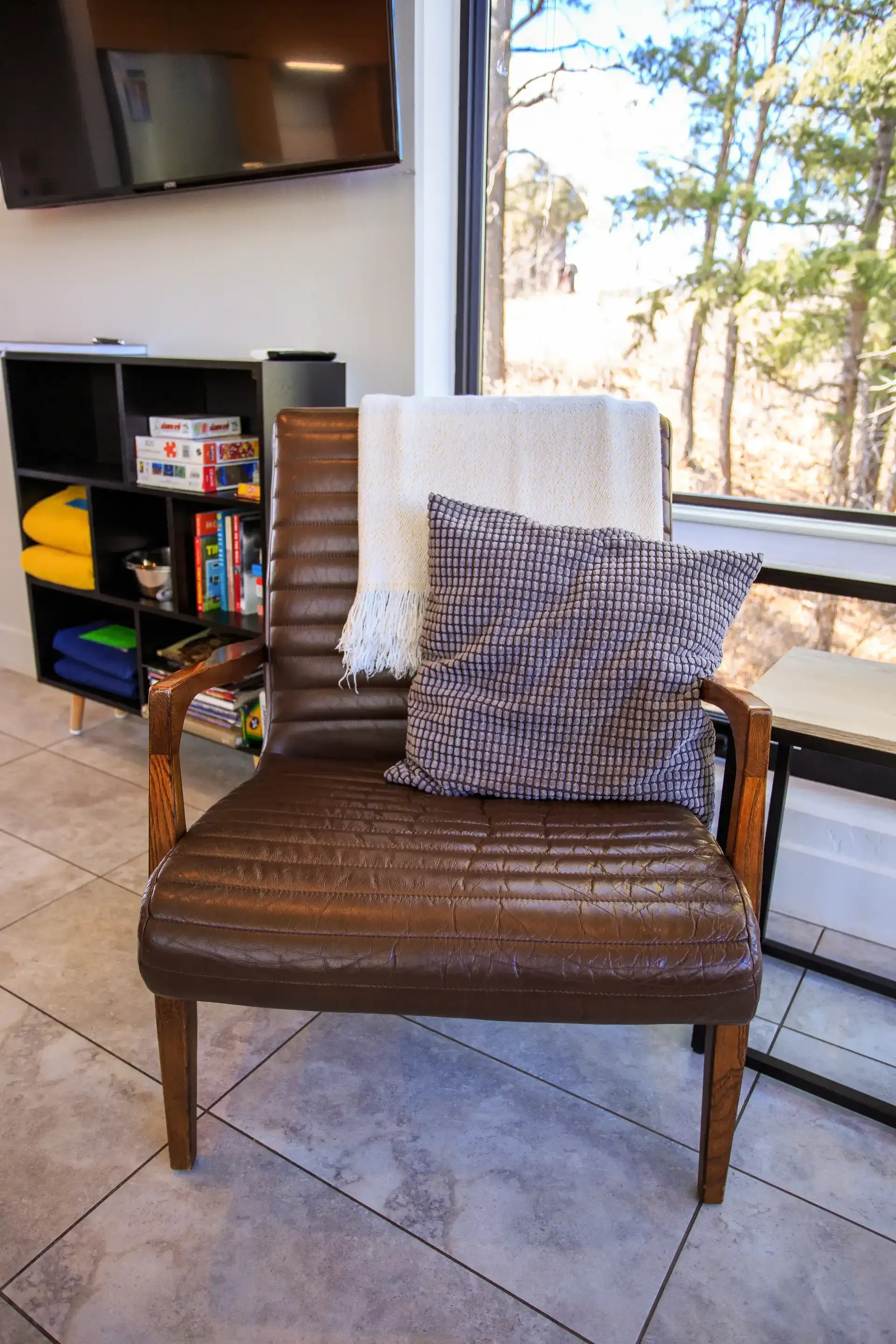 A brown, ribbed armchair with a white throw blanket and patterned throw pillow sits on a tiled floor by a window.