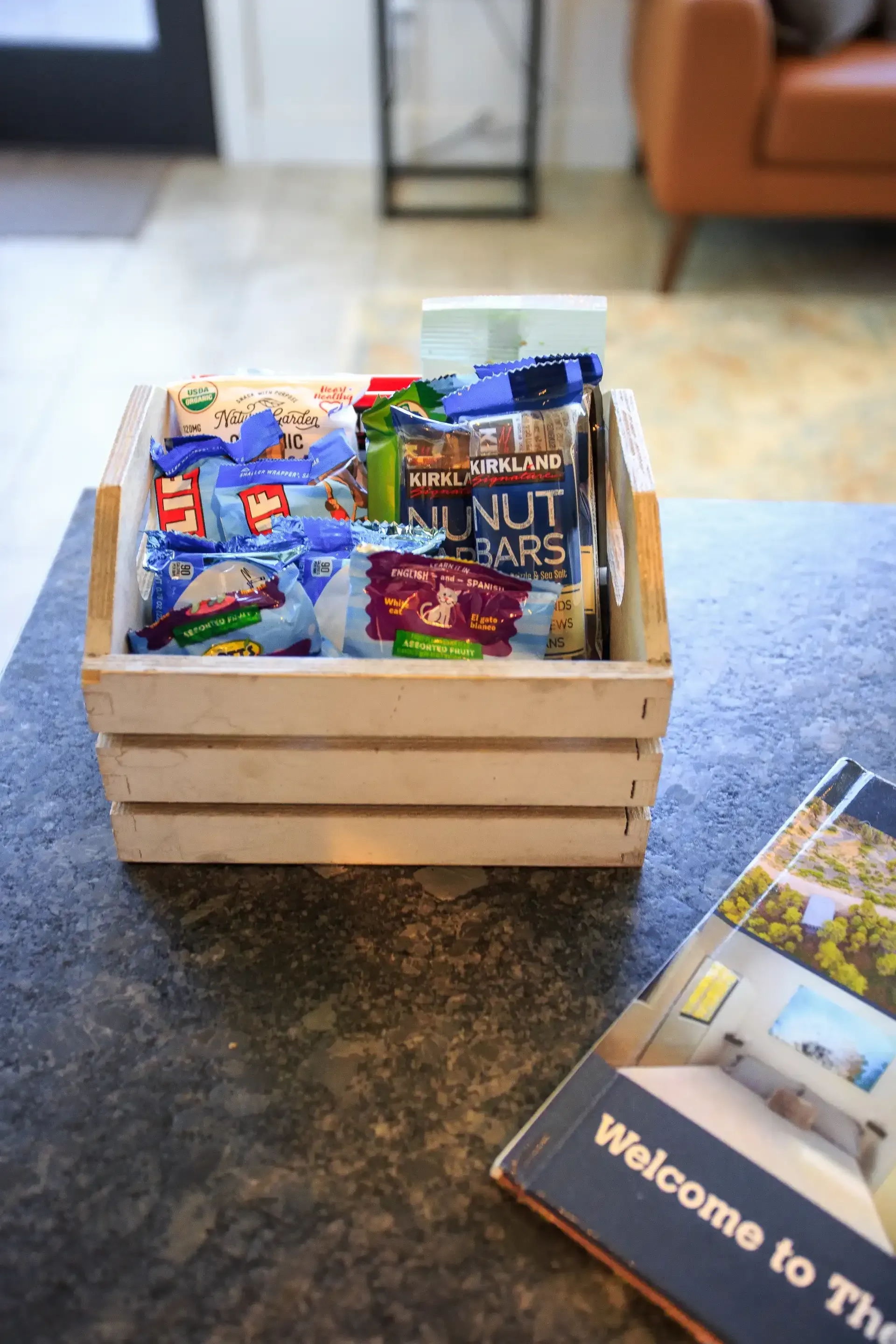 A wooden crate filled with various packaged snacks sits on a counter next to a welcome brochure.
