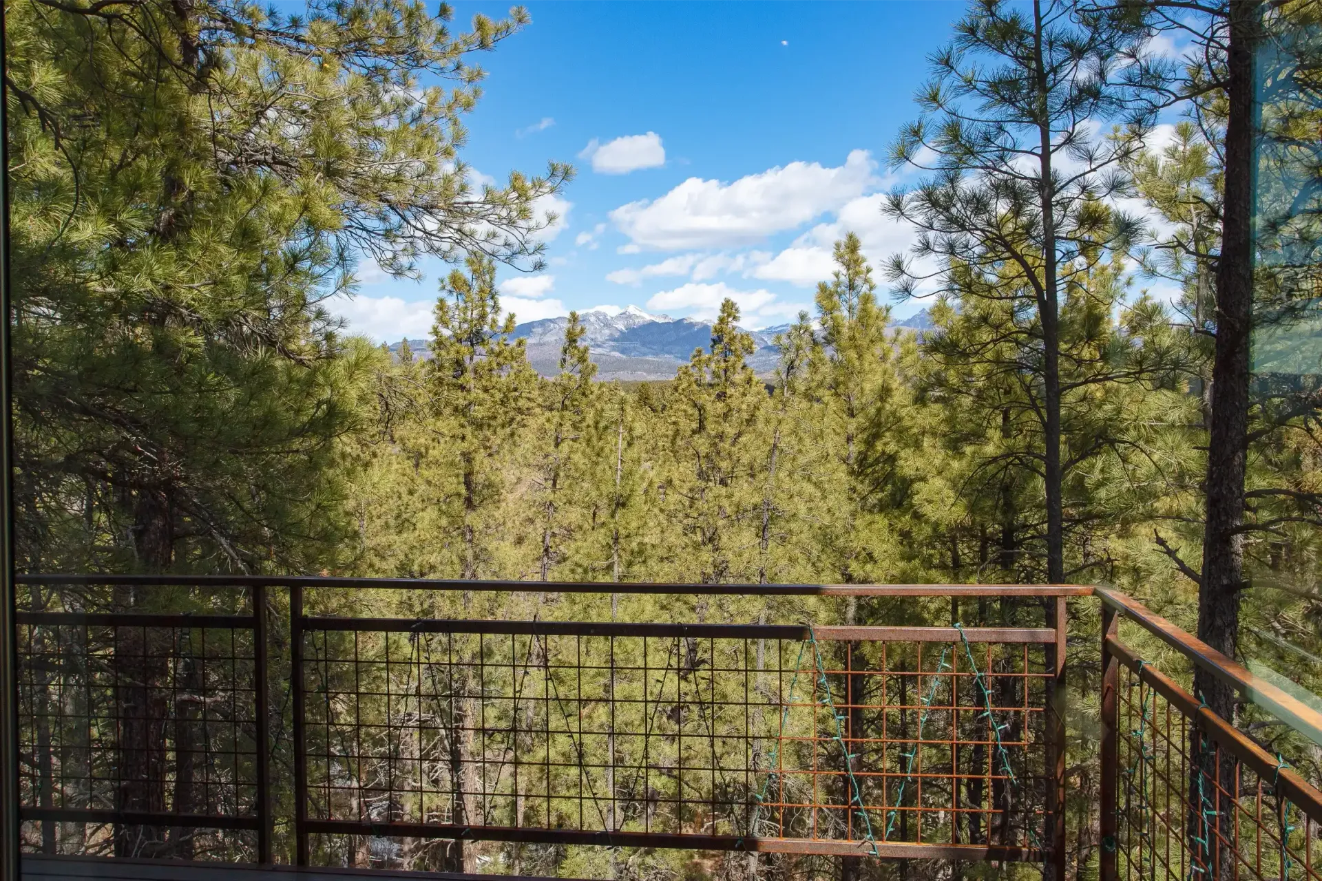 A metal balcony railing overlooks a dense forest of evergreen trees under a blue, partly cloudy sky with distant mountains.