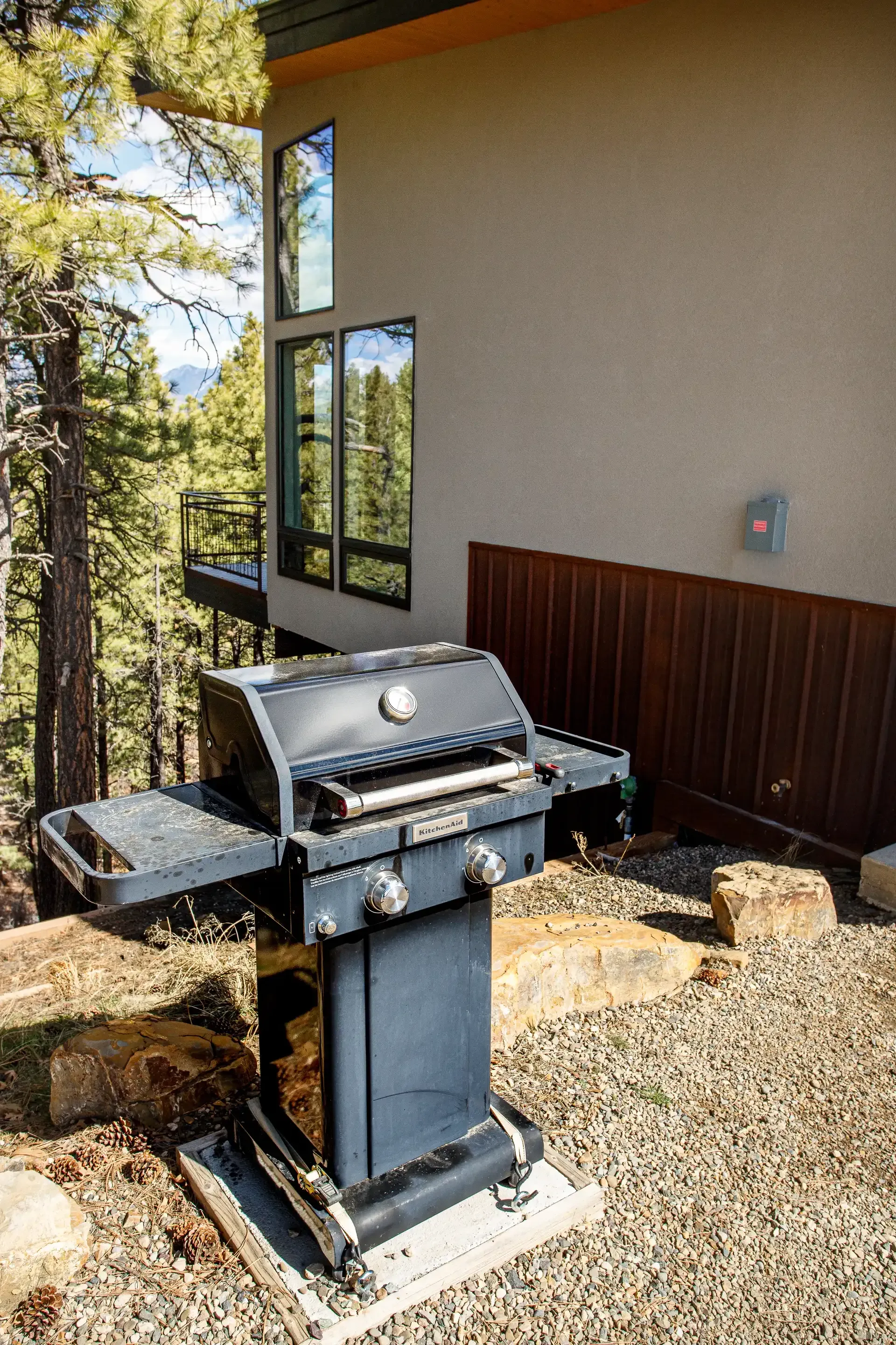 A dark gray barbecue grill stands on a gravel patio outside a house with brown siding and windows overlooking a forest.