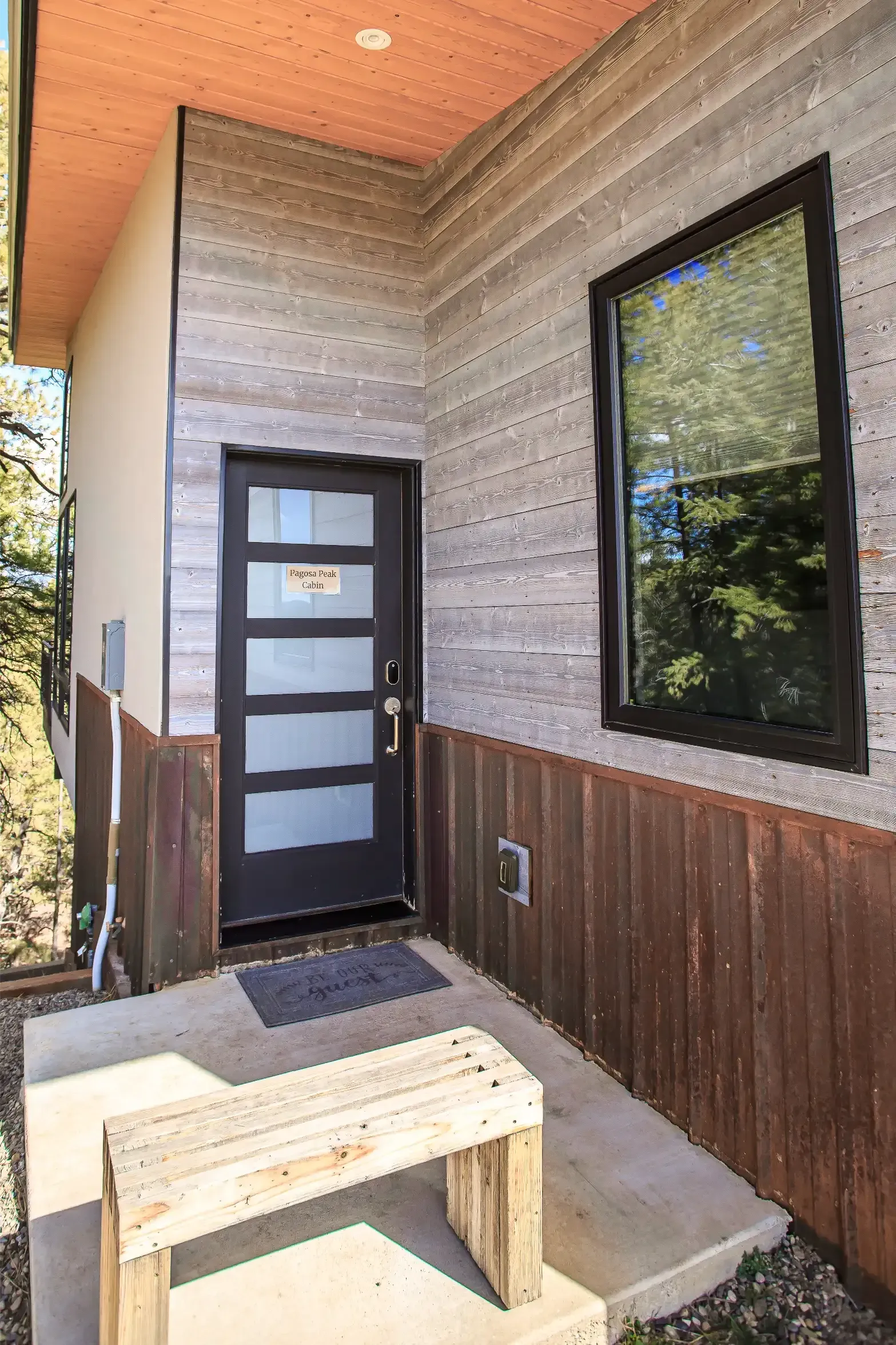 A modern front entrance with a dark door, light wood walls, rustic metal siding, and a wooden bench on a concrete stoop.