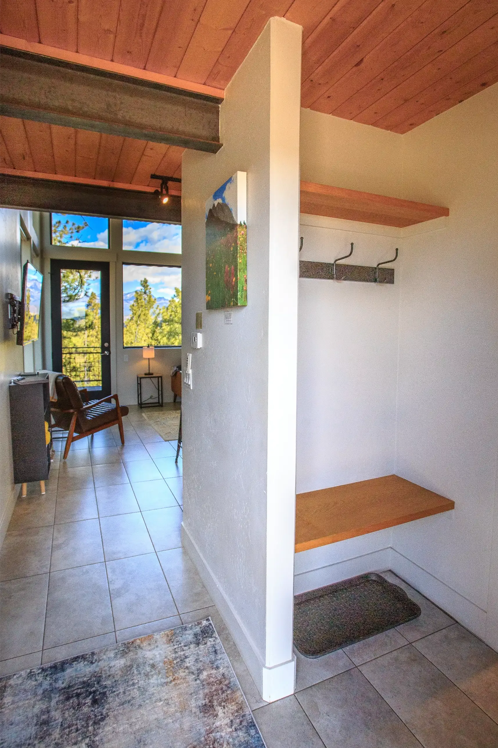 An entryway with a wooden bench, coat hooks, and a half-wall partition looking into a sunlit living room with tiled floors.