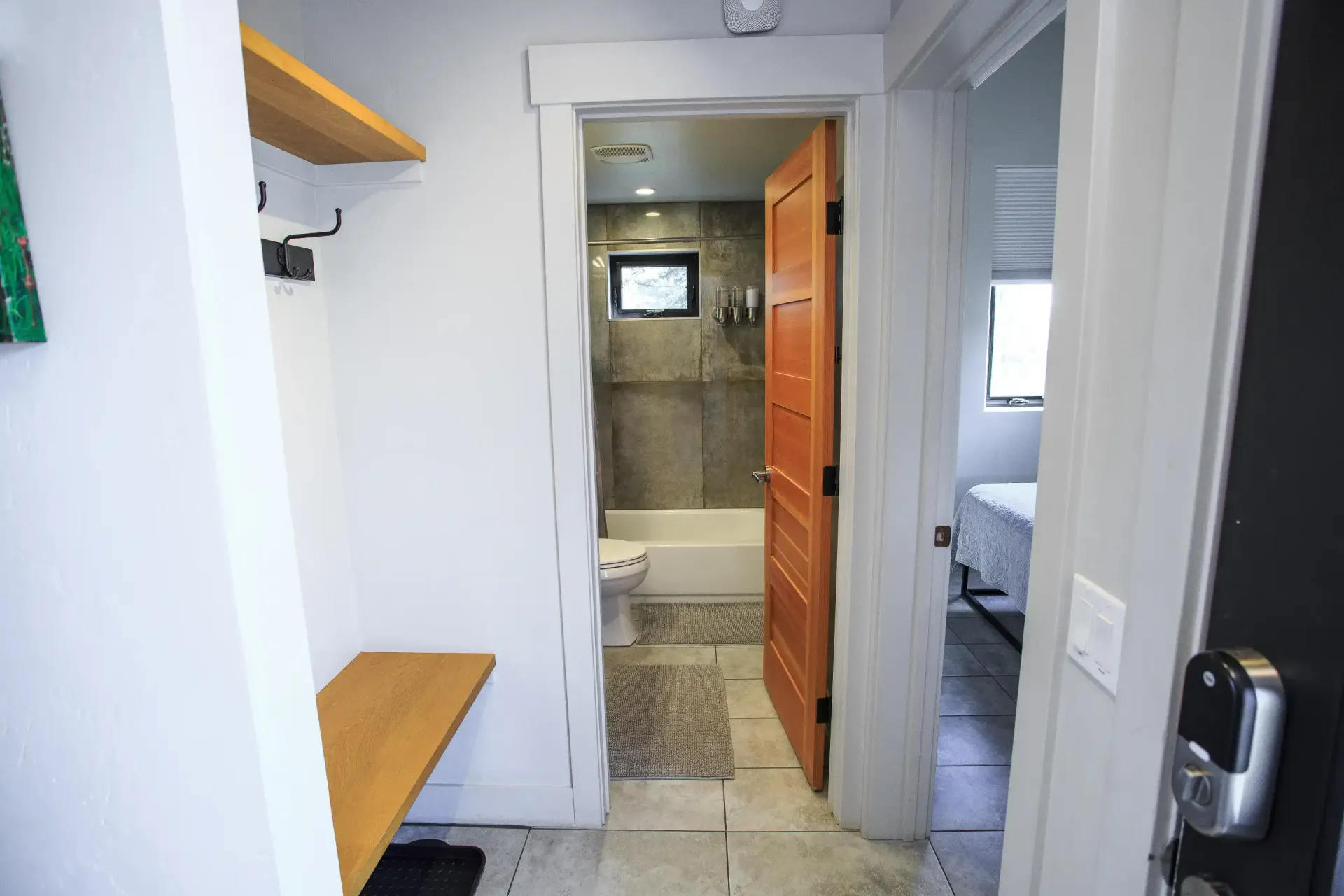 A hallway with a wooden coat rack and bench leads to a bathroom with a tub, with a bedroom visible through a side doorway.