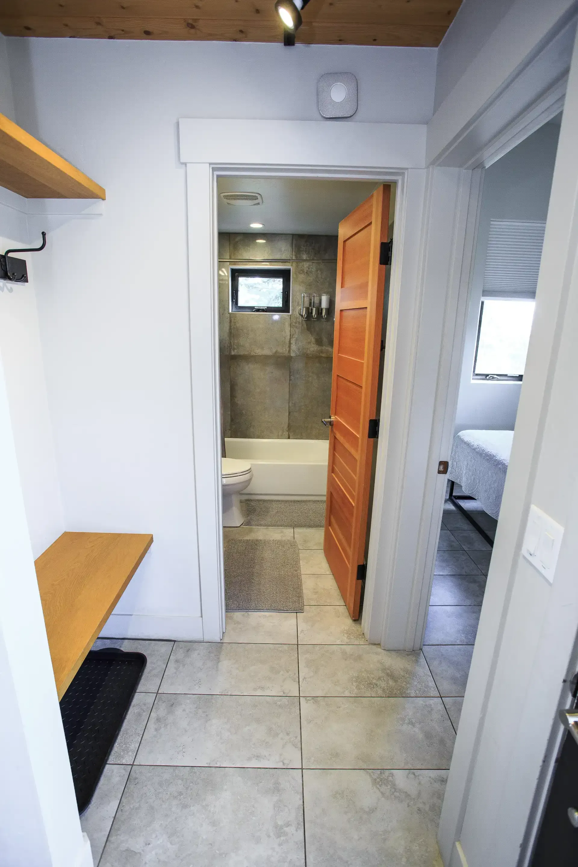 A hallway with a wooden bench and wall hook leads to a bathroom with a tiled floor and a wooden door.