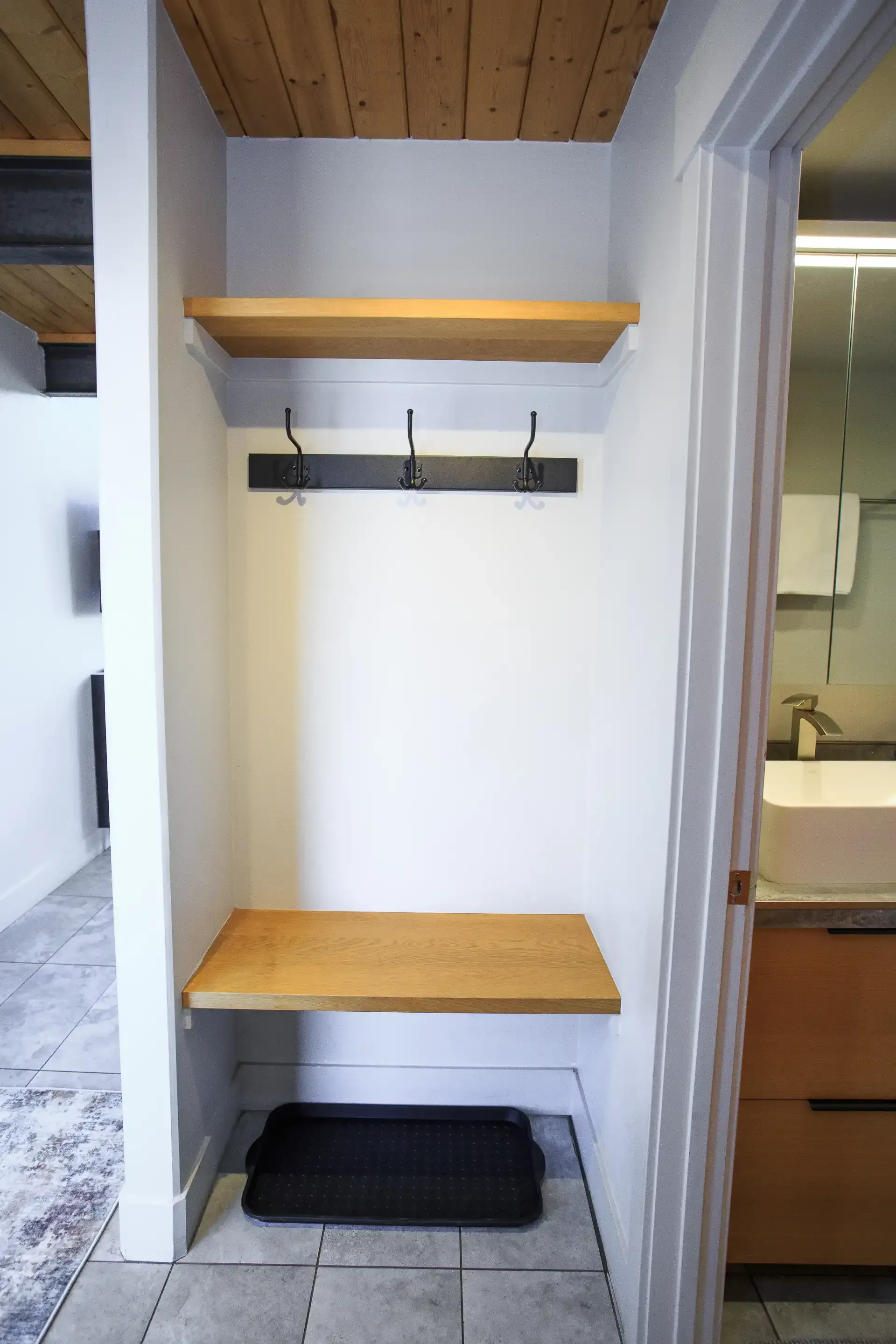 A hallway mudroom area with a wooden bench, a shelf above coat hooks, and a black shoe tray on a tiled floor.