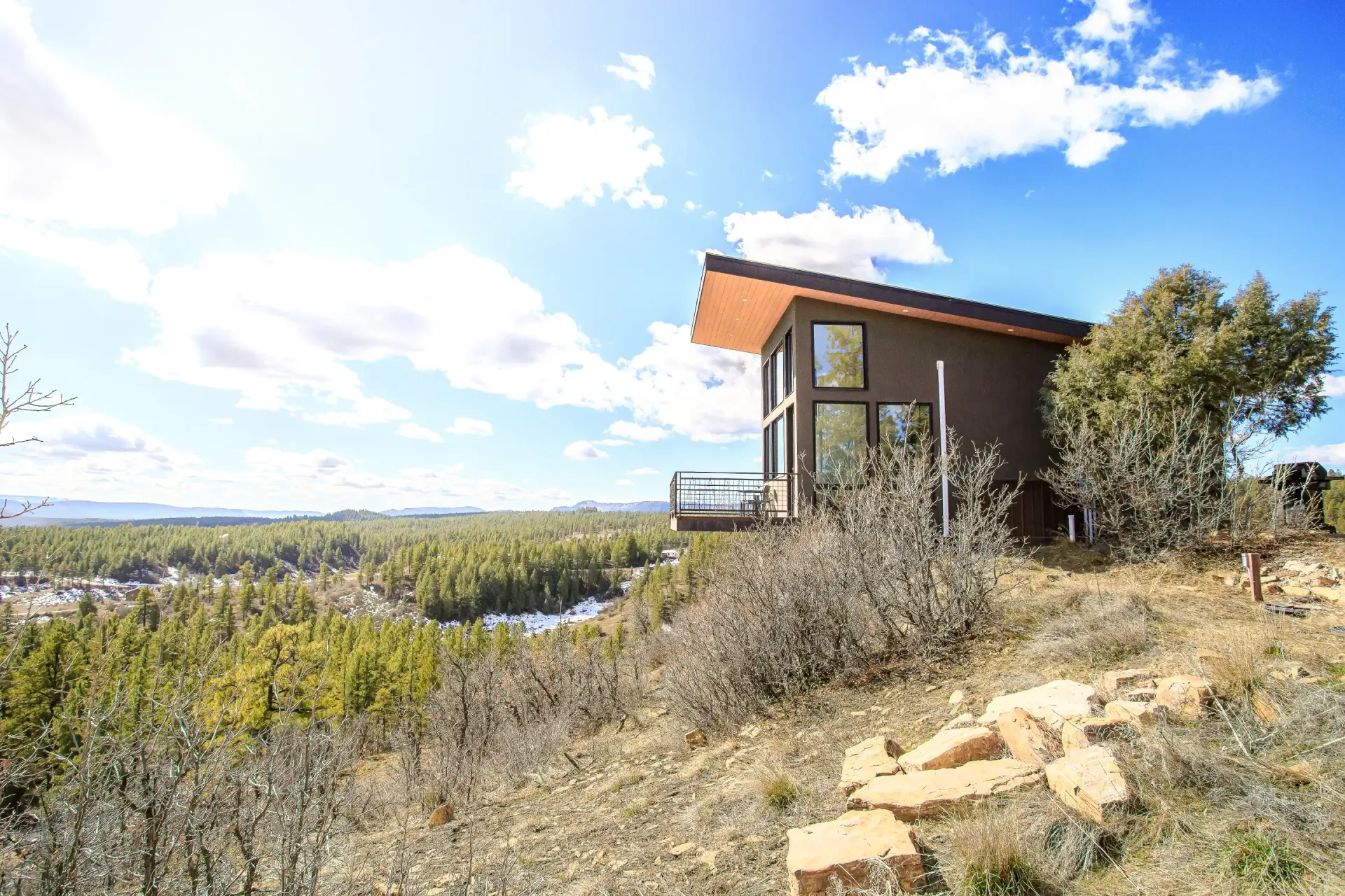 A modern, flat-roofed cabin perched on a hillside overlooking a vast pine forest under a bright, partly cloudy sky.