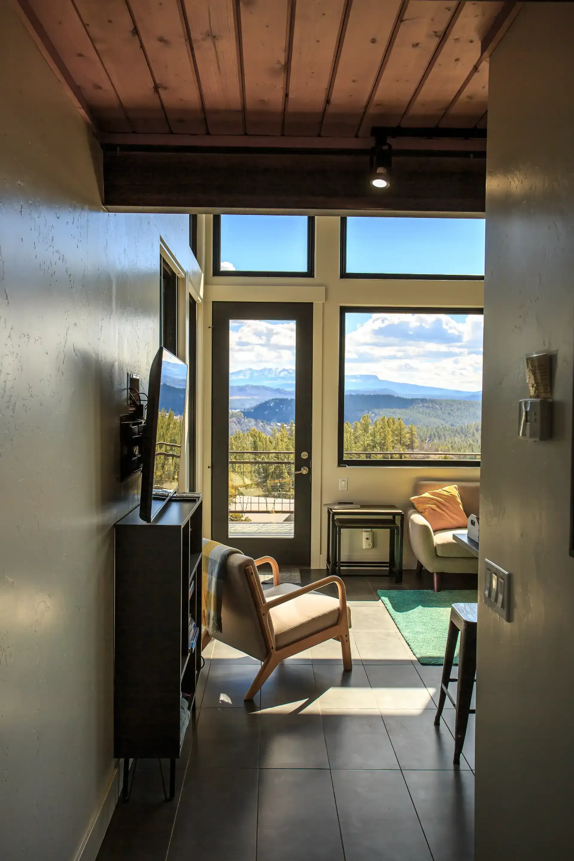 A view from a hallway into a living room with large windows, a glass door, a wooden ceiling, and a scenic mountain view.