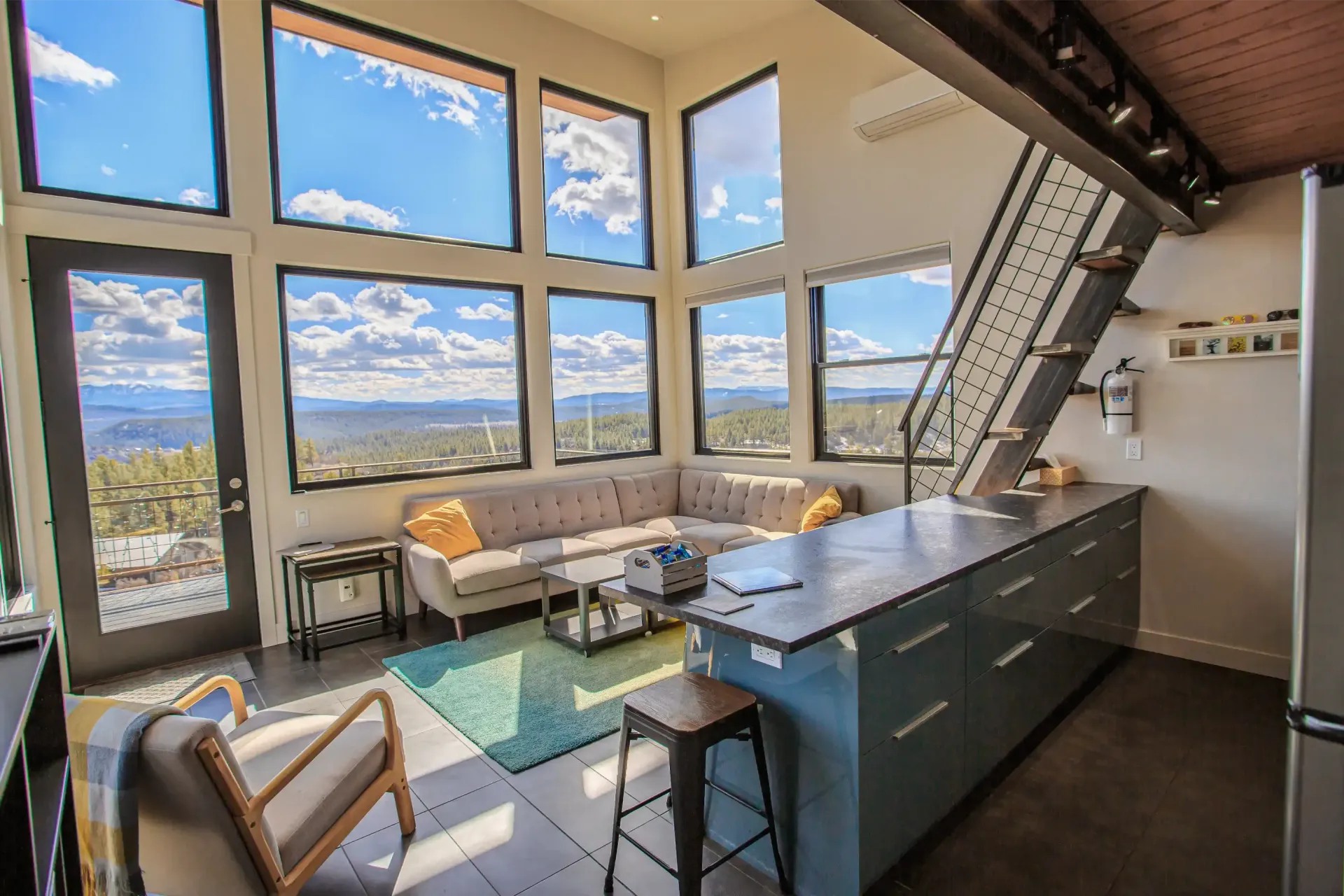 Modern living room with a sectional sofa, blue kitchen island, and tall windows showing a scenic, forested mountain view.