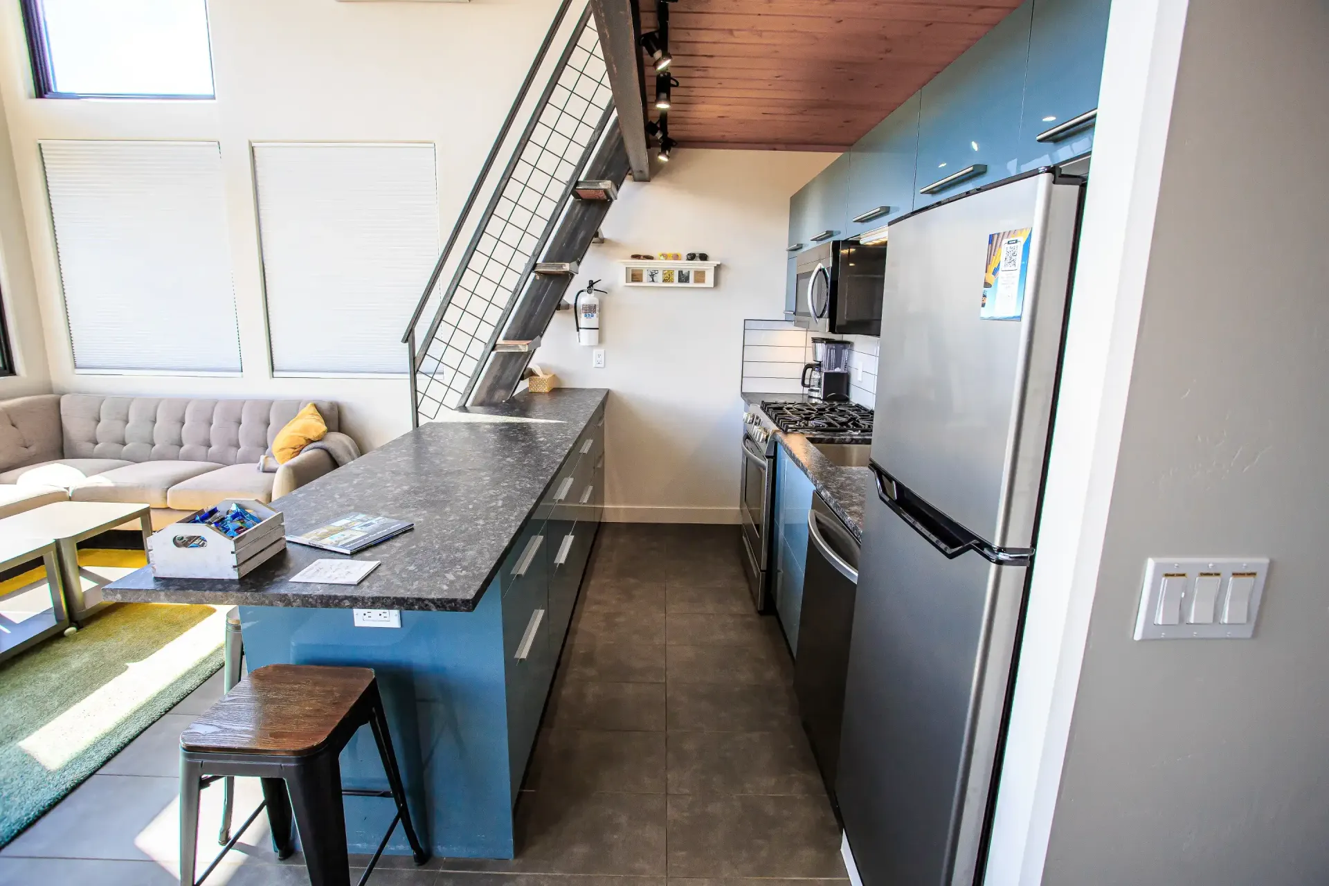 A view of a modern loft kitchen featuring a blue island, stainless steel refrigerator, and metal staircase.