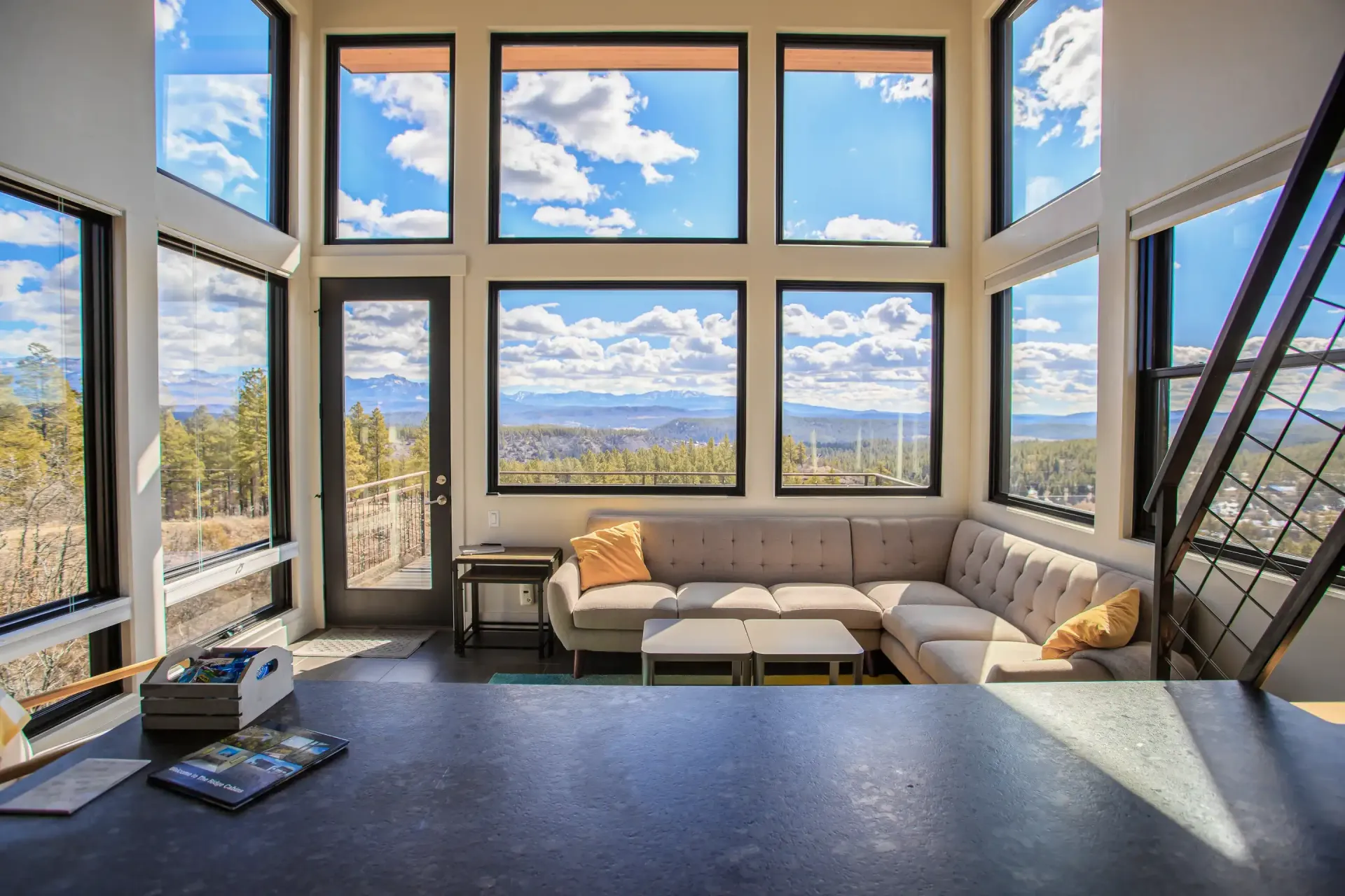 A light-filled living area featuring a tan sectional sofa, glass walls with outdoor views, and a dark kitchen countertop.