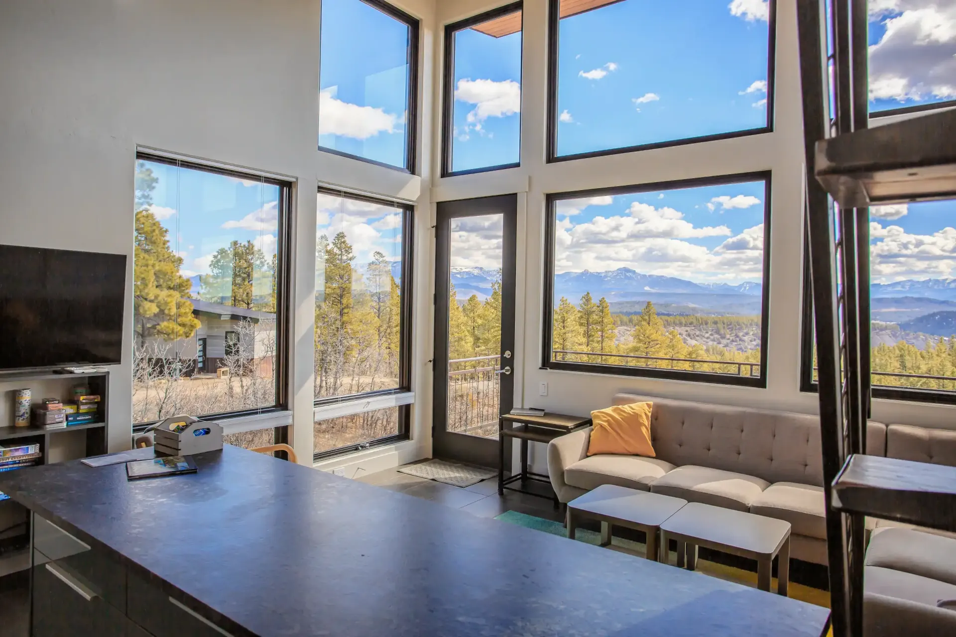 A modern, high-ceilinged living area with floor-to-ceiling windows, a sofa, and a dark kitchen island overlooking mountains.