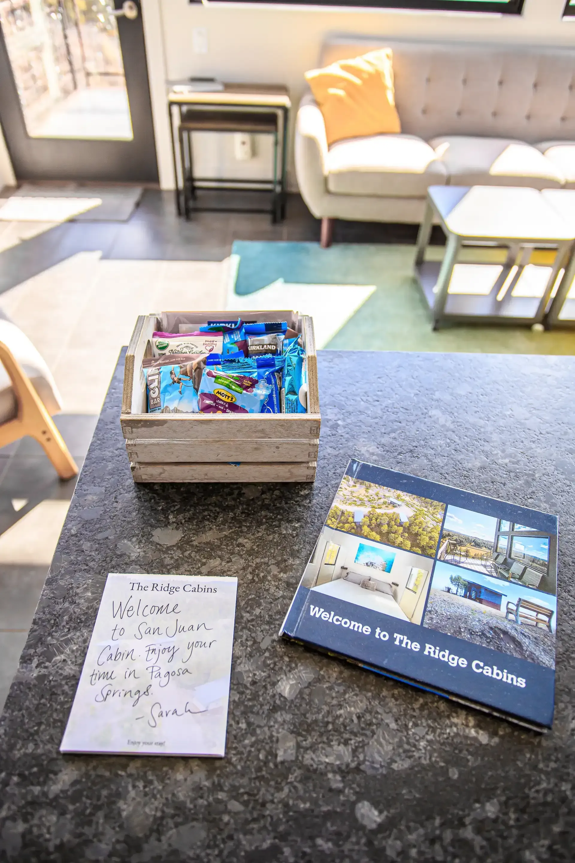 A granite countertop holds a wooden crate of snacks, a handwritten note, and a 