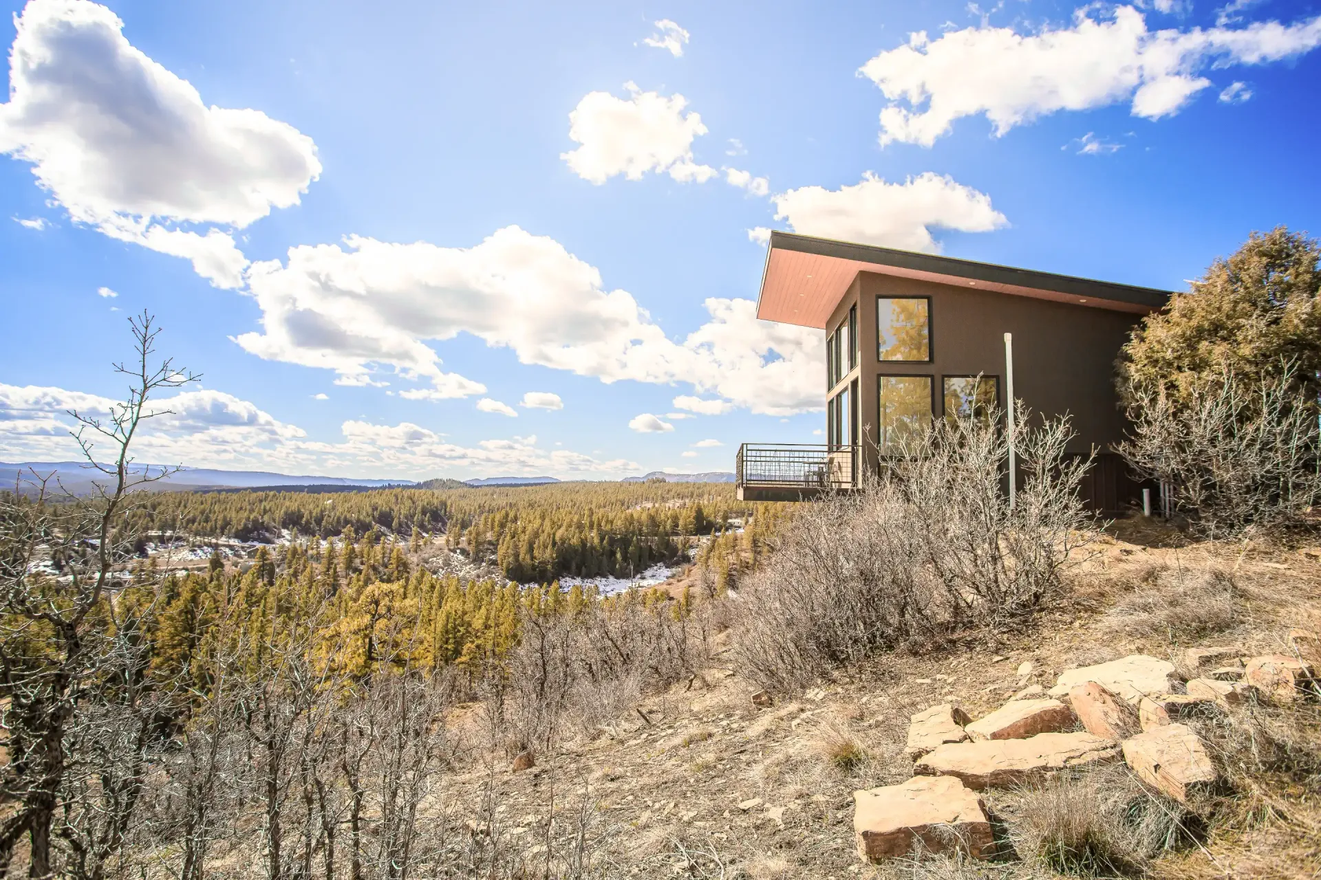 A modern cabin with a slanted roof overlooks a vast forest valley under a bright blue, partly cloudy sky.