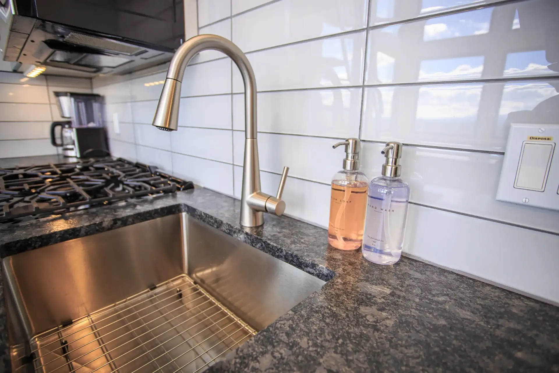 A modern kitchen sink with a stainless steel faucet, subway tile backsplash, and two soap dispensers on dark granite.