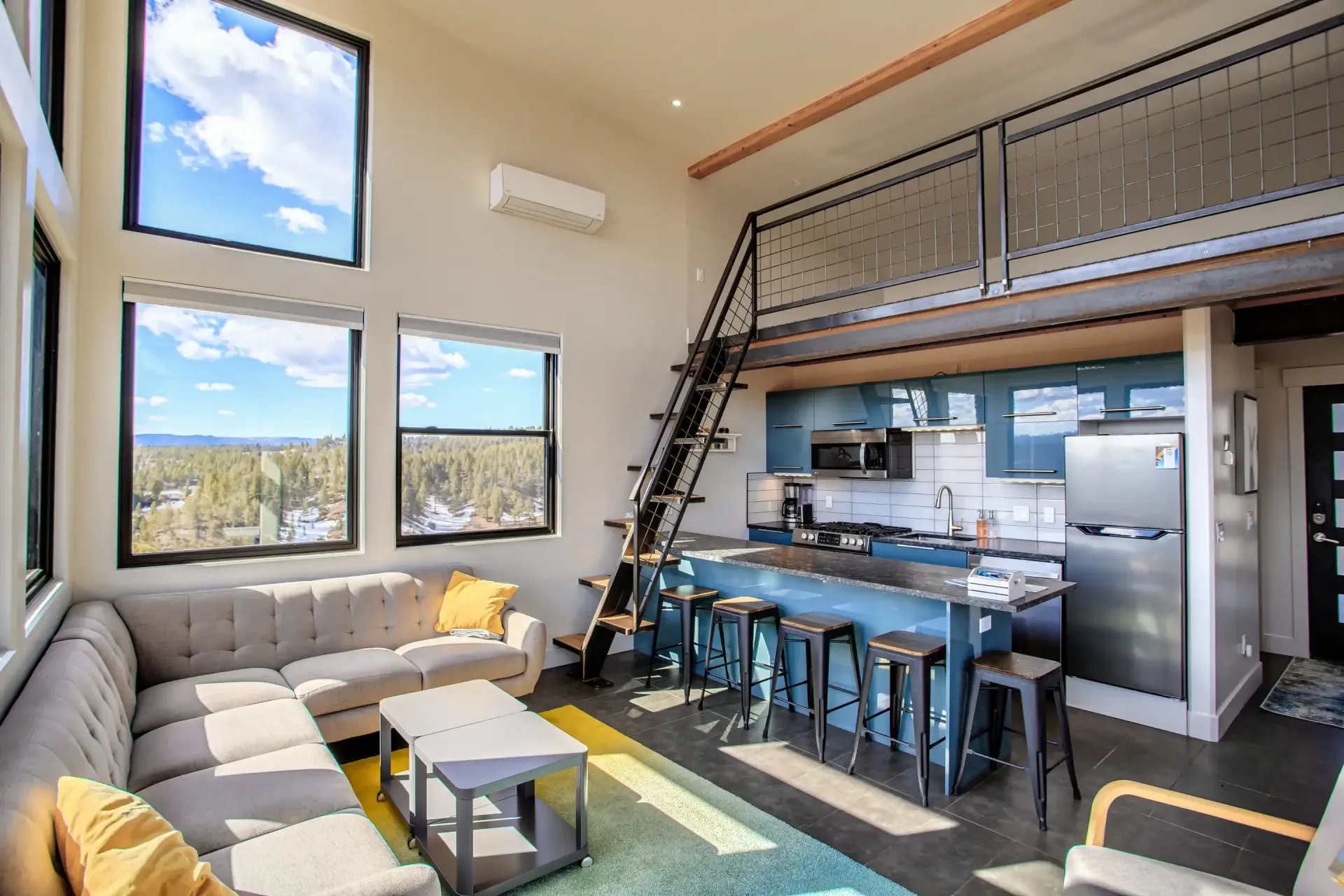 Modern tiny home living area featuring a gray sectional sofa, yellow accents, a kitchen island, and a loft bedroom above.