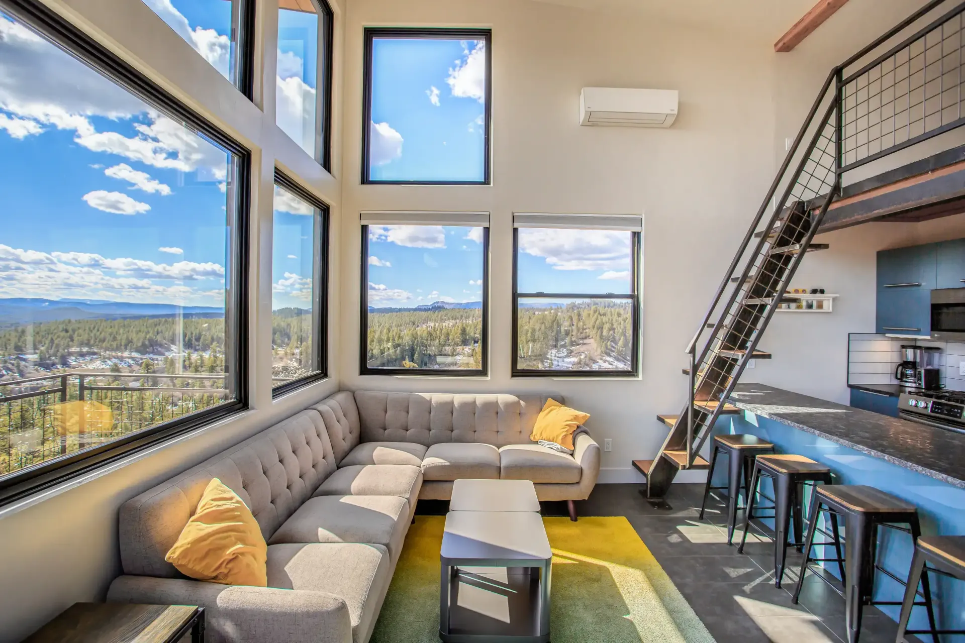 A modern living room with a gray sectional sofa, yellow accents, and large windows offering a panoramic forest view.